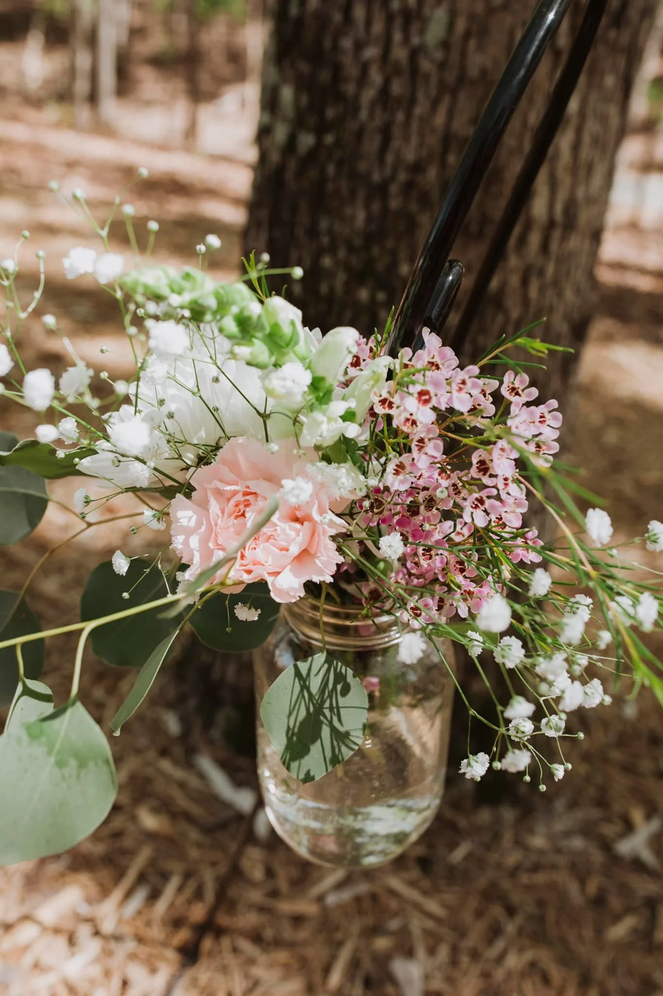 A glass jar filled with pink, white, and green flowers, including small white baby's breath, set on the ground against a tree trunk in a natural outdoor setting.