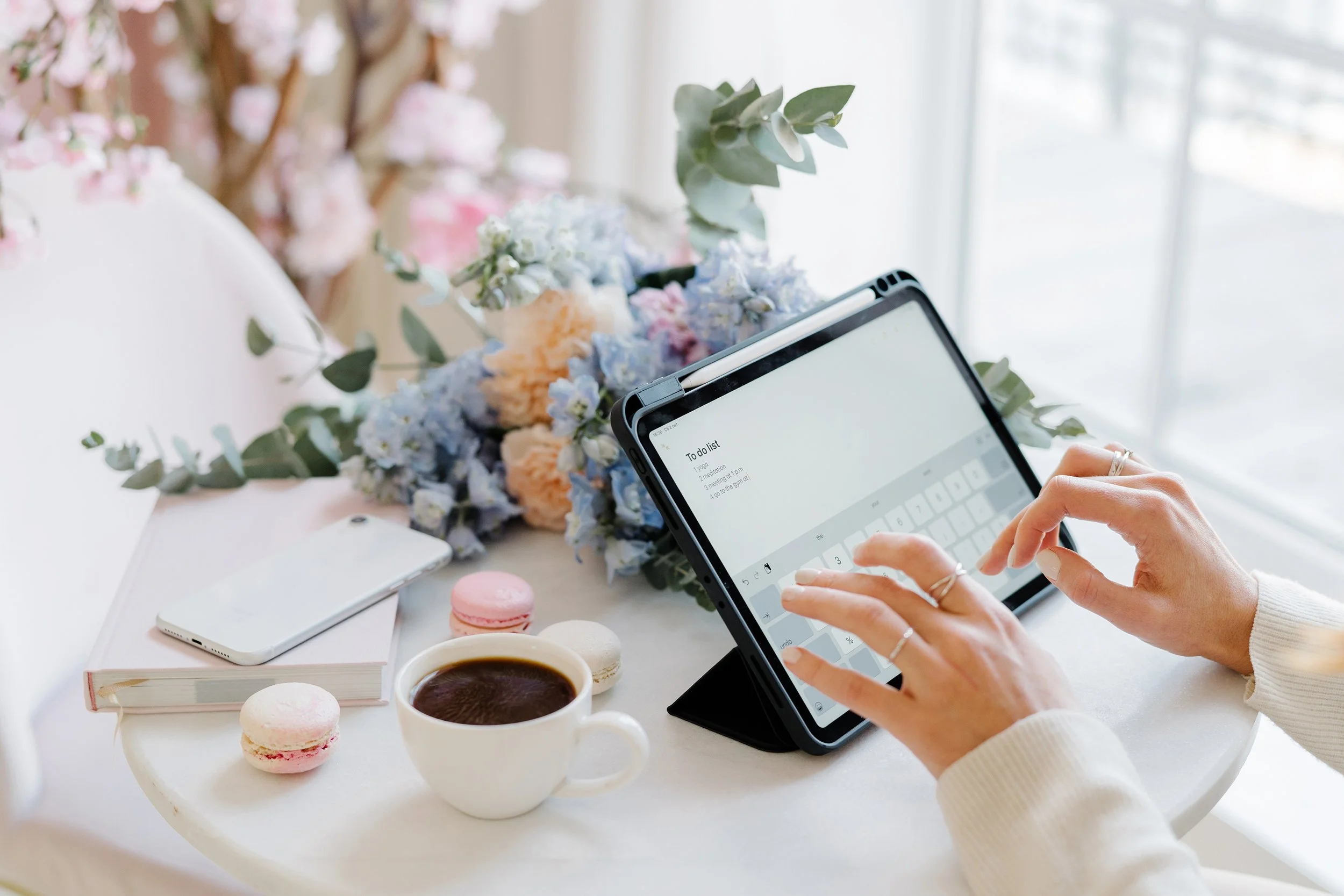 Person using a tablet with a to-do list on a table surrounded by flowers, macarons, a cup of coffee, and a smartphone.