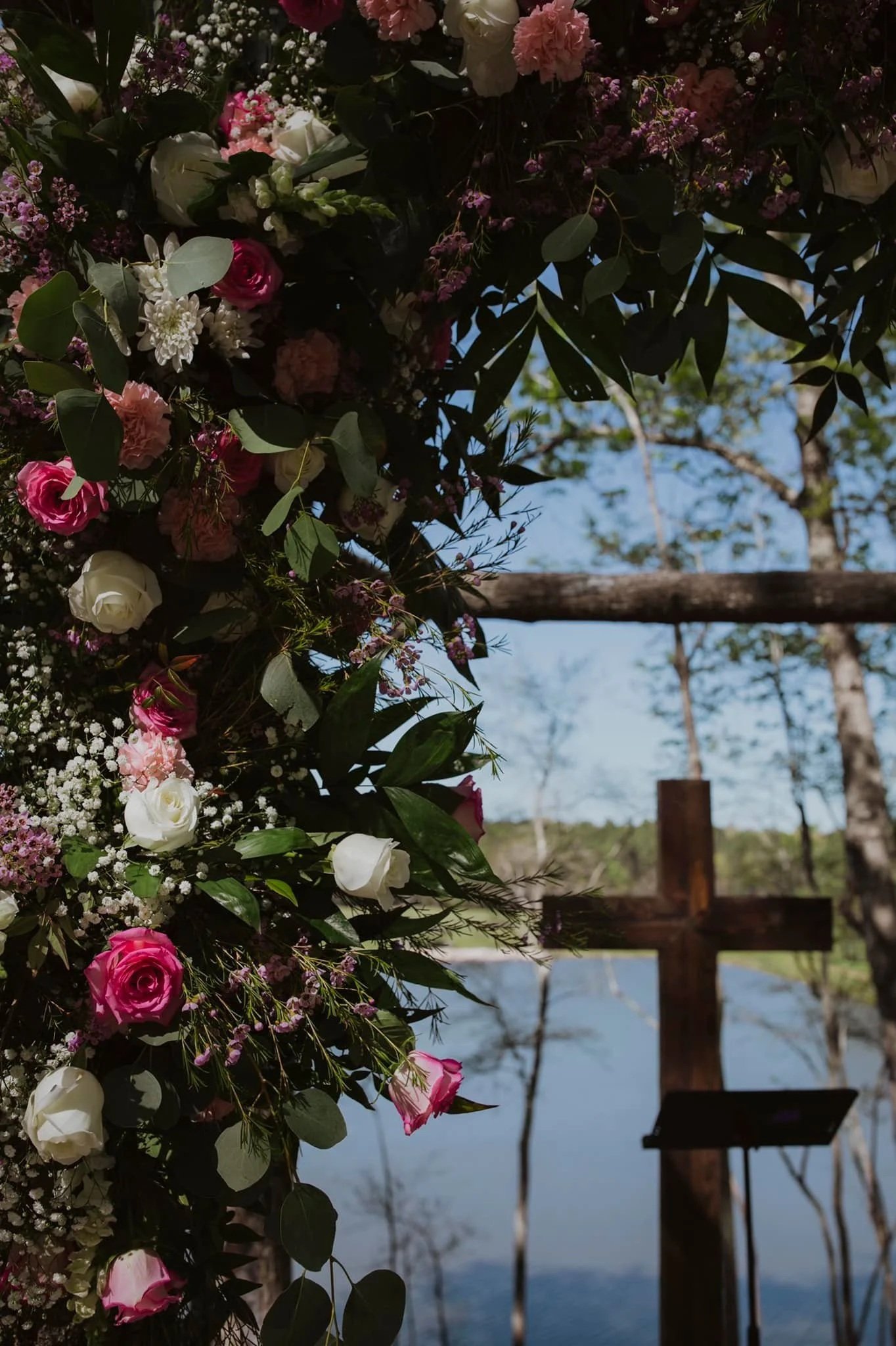 Close-up view of a floral arrangement with pink, white, and purple flowers along with green leaves, set outdoors with a wooden cross and a lake in the background.