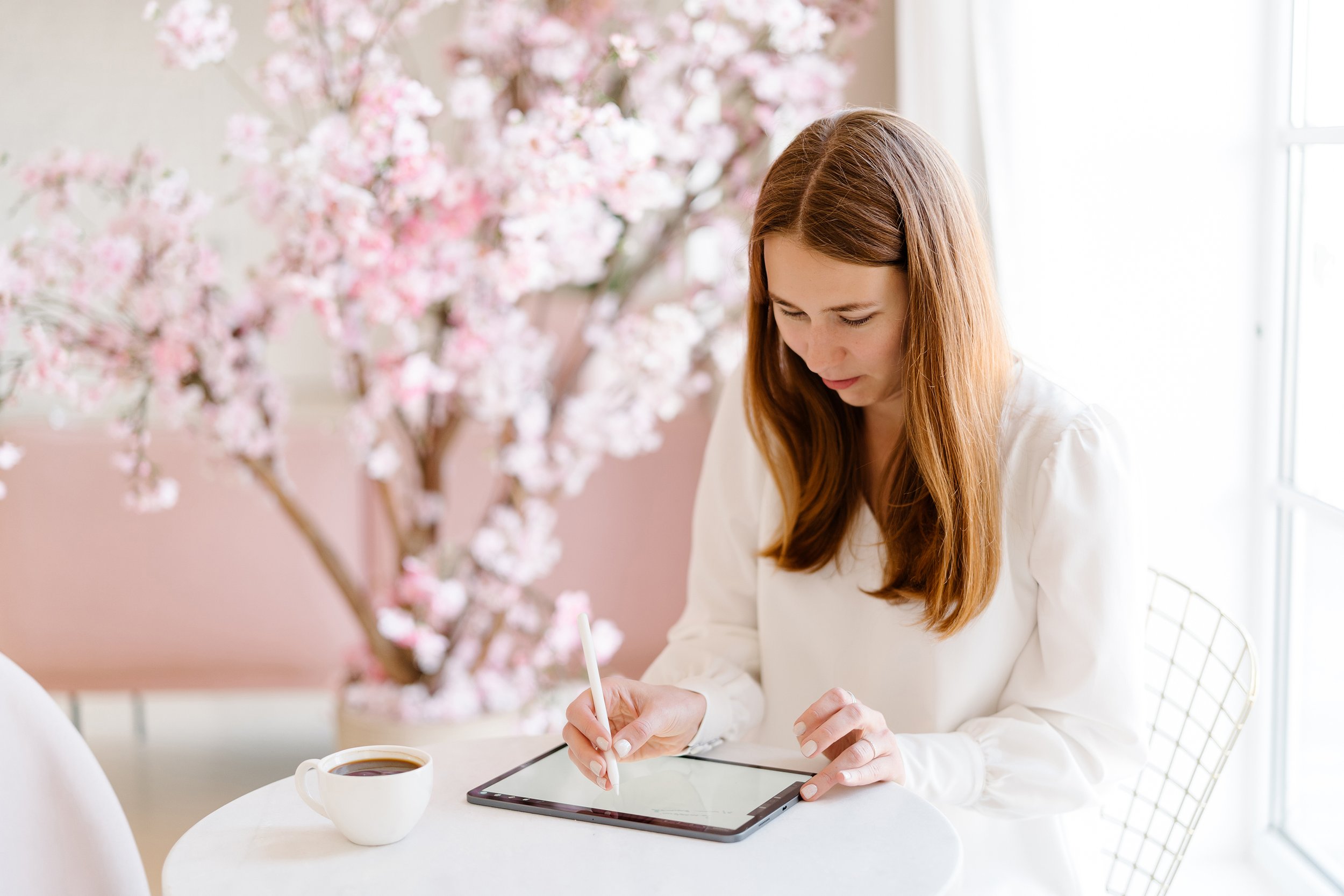 A woman with long red hair using a stylus on a tablet, sitting at a white table with a cup of coffee near a pink blossom tree in a bright, indoor setting.