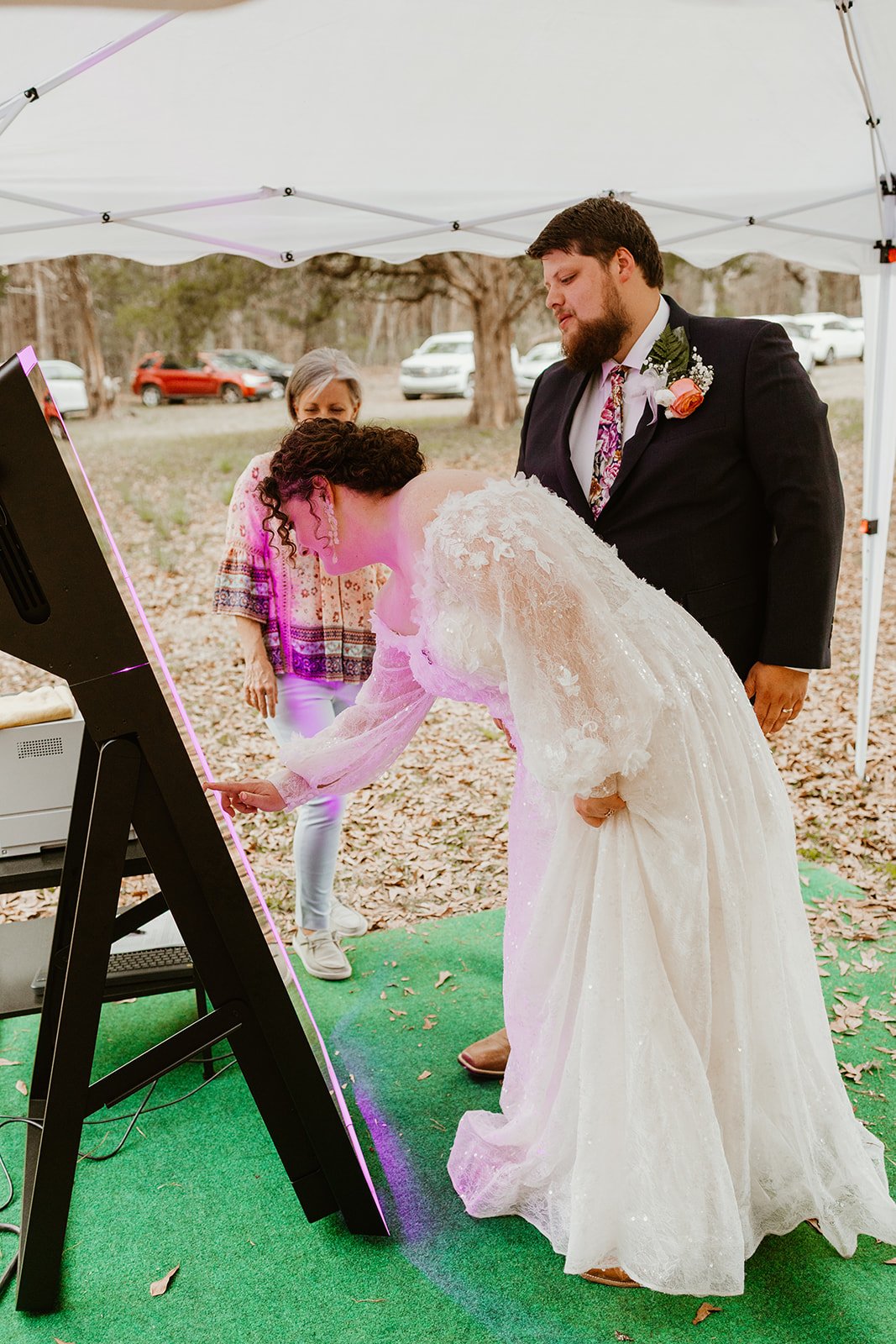 Bride and groom at their wedding, lighting a unity candle, with a woman standing behind them and a green carpet on the ground, outdoors under a white canopy.