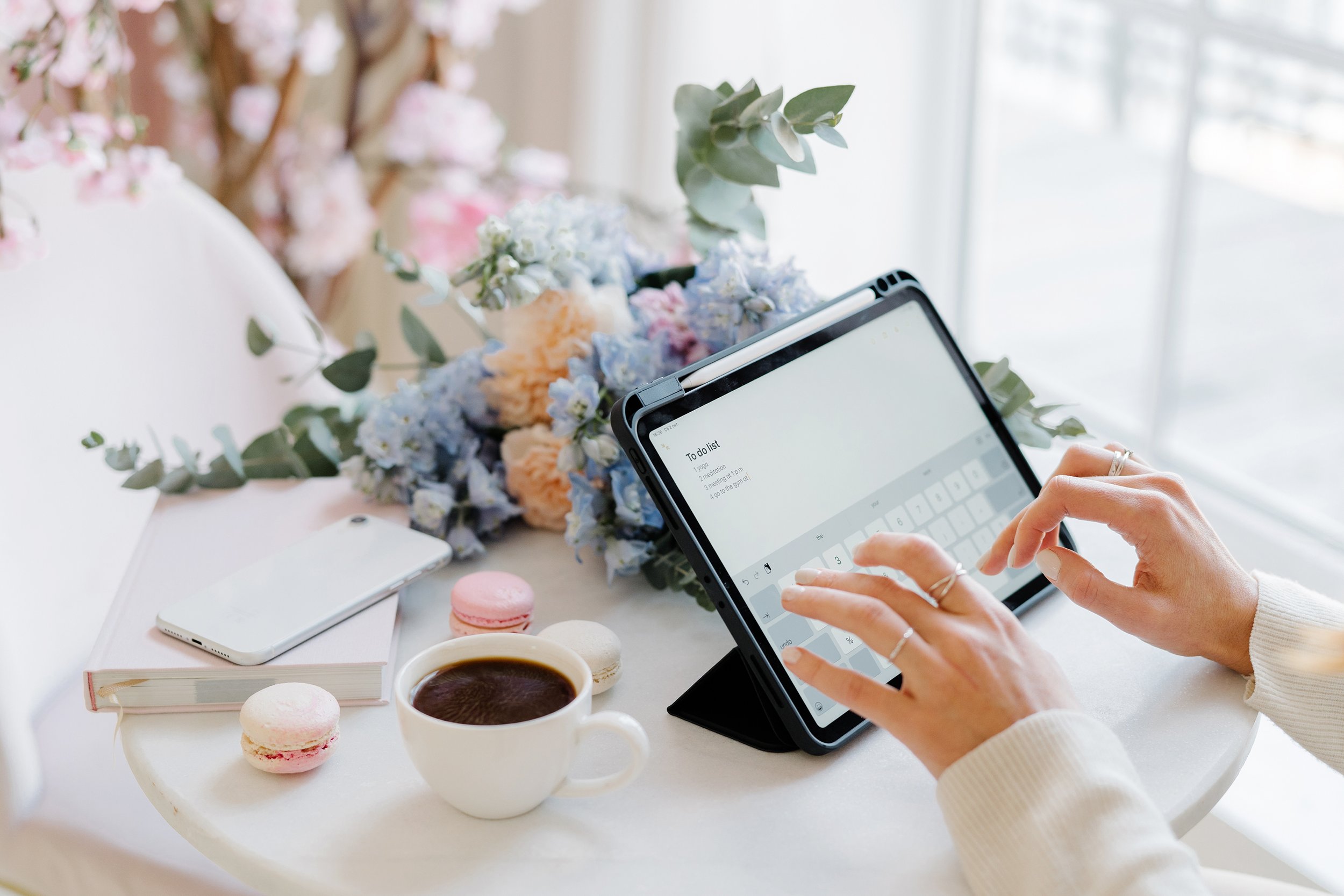 A person typing on a tablet with a to-do list, surrounded by a cup of coffee, macaron cookies, a smartphone, a book, and floral decorations on a white table.