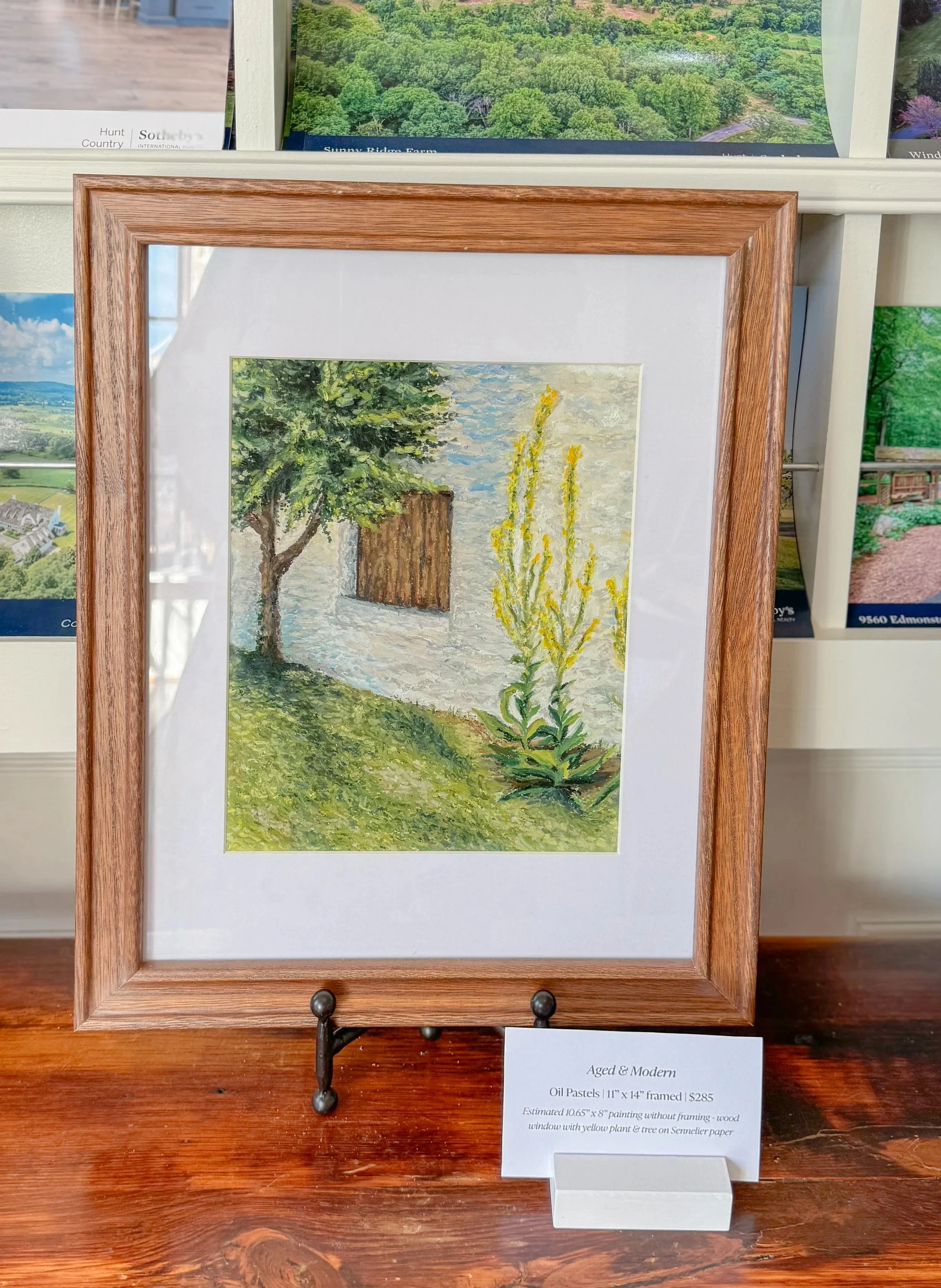 Framed oil pastel painting of a house with a tree and yellow flowers in front, placed on a wooden table.