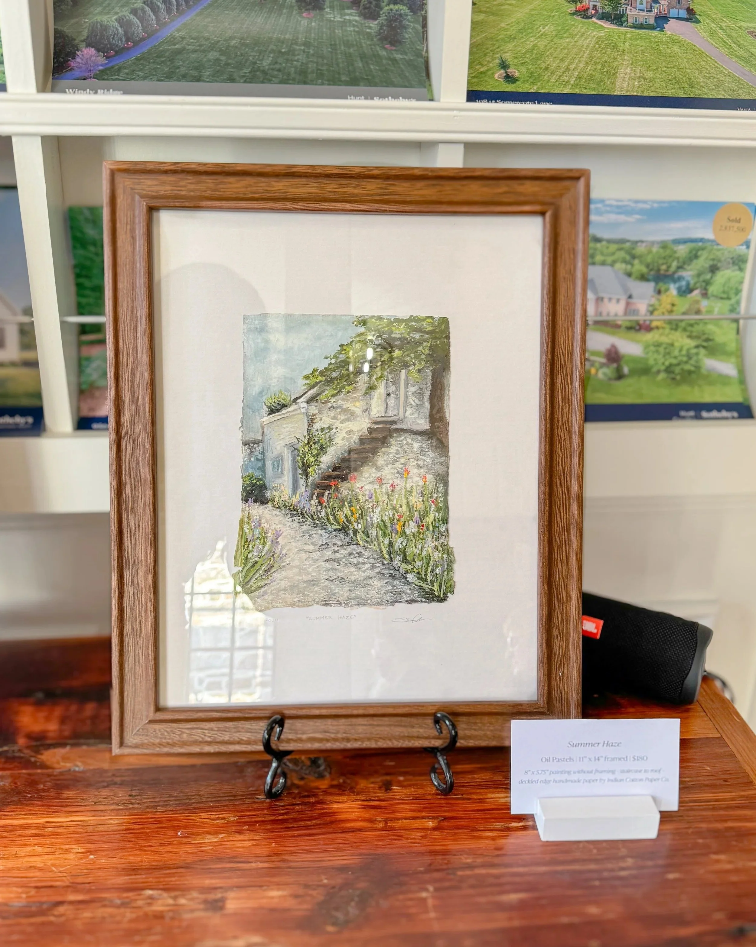 Oil pastel painting of a stone pathway leading to a white house with steps and a balcony, surrounded by greenery and flowers, titled 'Summer Haze' and framed in wood.