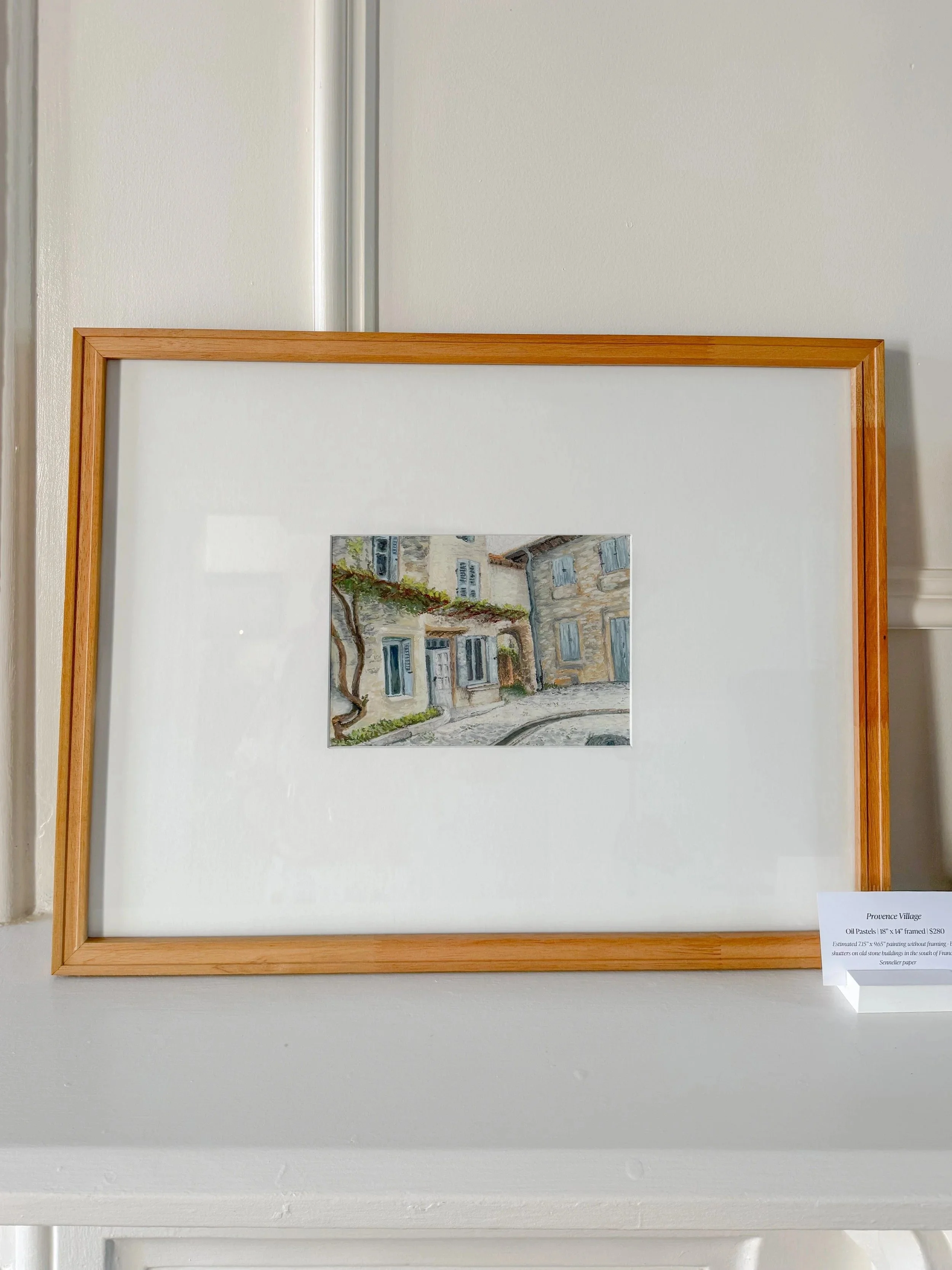 Small painting of a street scene with houses and a vine-covered building, displayed in a wooden frame on a white wall.