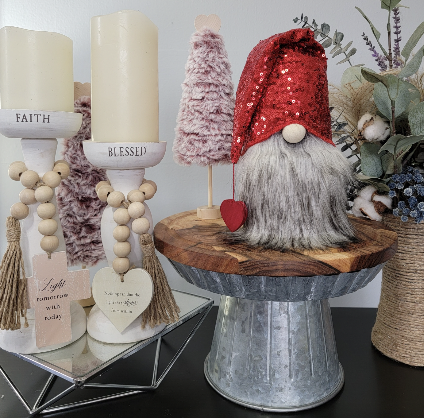 Decorative holiday display featuring two white candle holders with wood bead accents labeled 'FAITH' and 'BLESSED', pink faux fur trees, a gnome with a red sequin hat and a heart ornament, and a large basket with eucalyptus and cotton bushes.