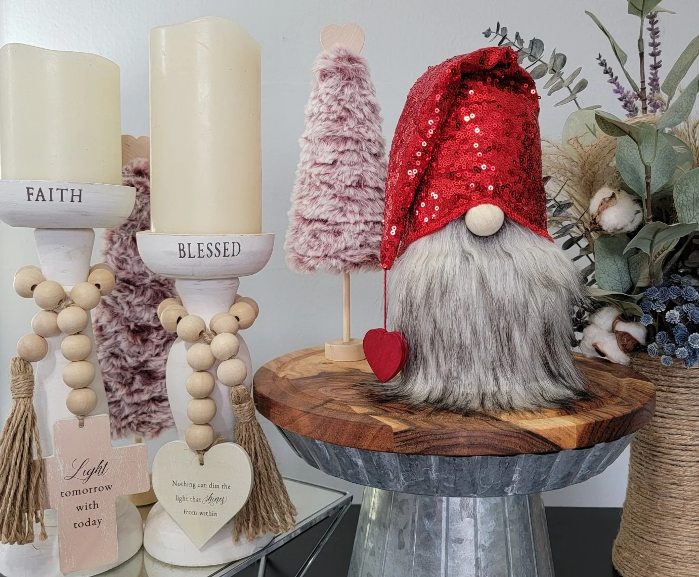 Decorative display with candles labeled 'FAITH' and 'BLESSED', a pink fluffy tree, a gnome figurine with a long gray beard, red sequin hat with a wooden nose, sitting on a wooden table, and greenery with purple and white flowers in the background.