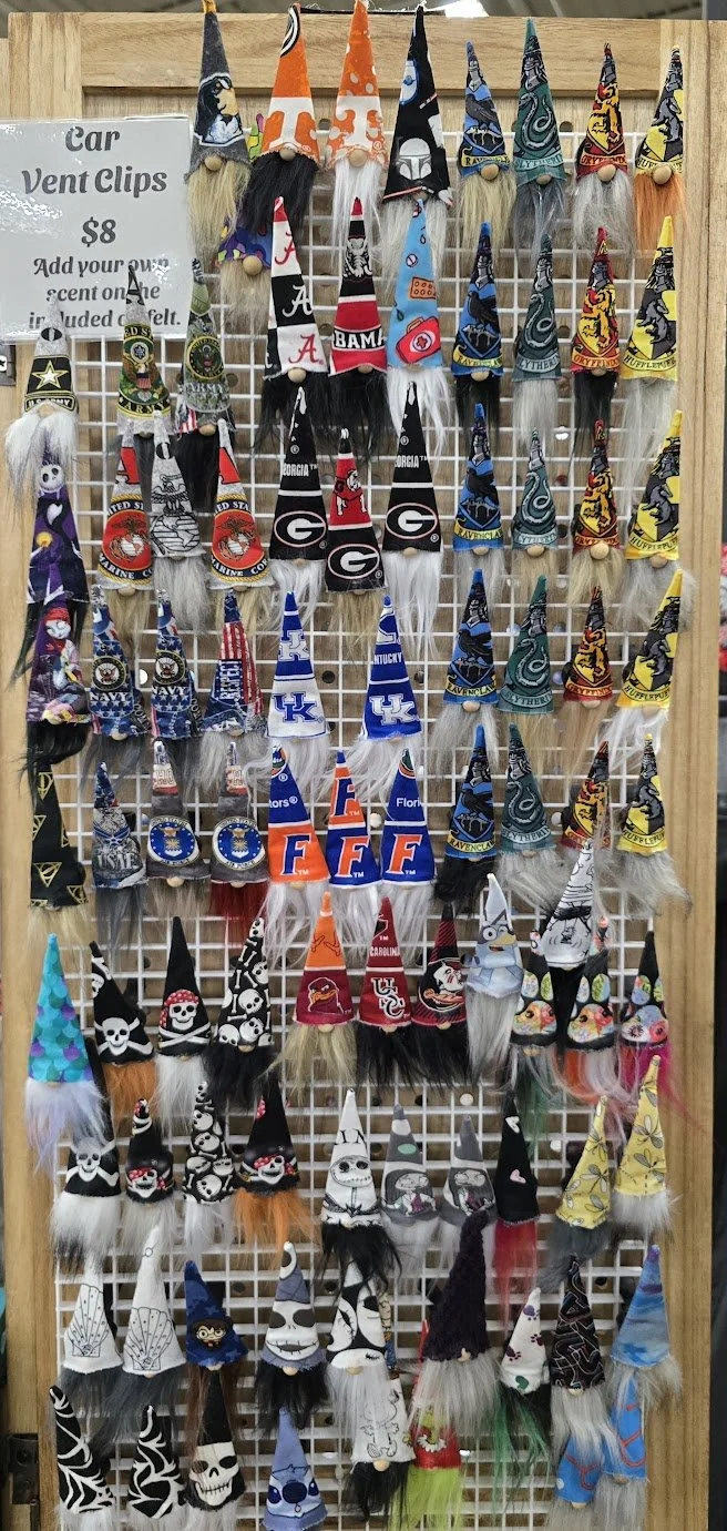 A display board with numerous car vent clips shaped like gnomes, each wearing hats with various sports team logos, characters, or themed decorations.