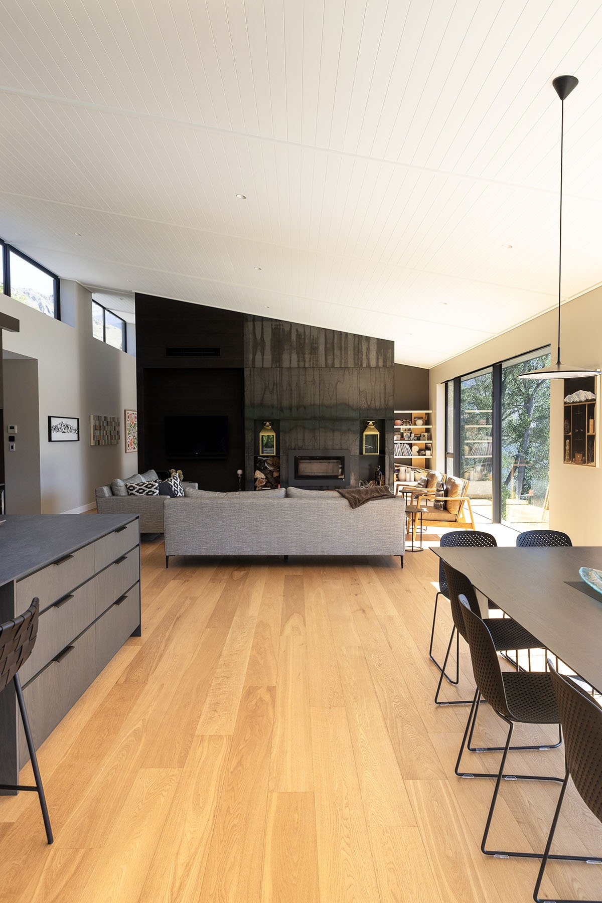 Modern living room with hardwood floors, gray sofa, black fireplace, built-in bookshelves, large windows with views of trees, and a dining table with black chairs.