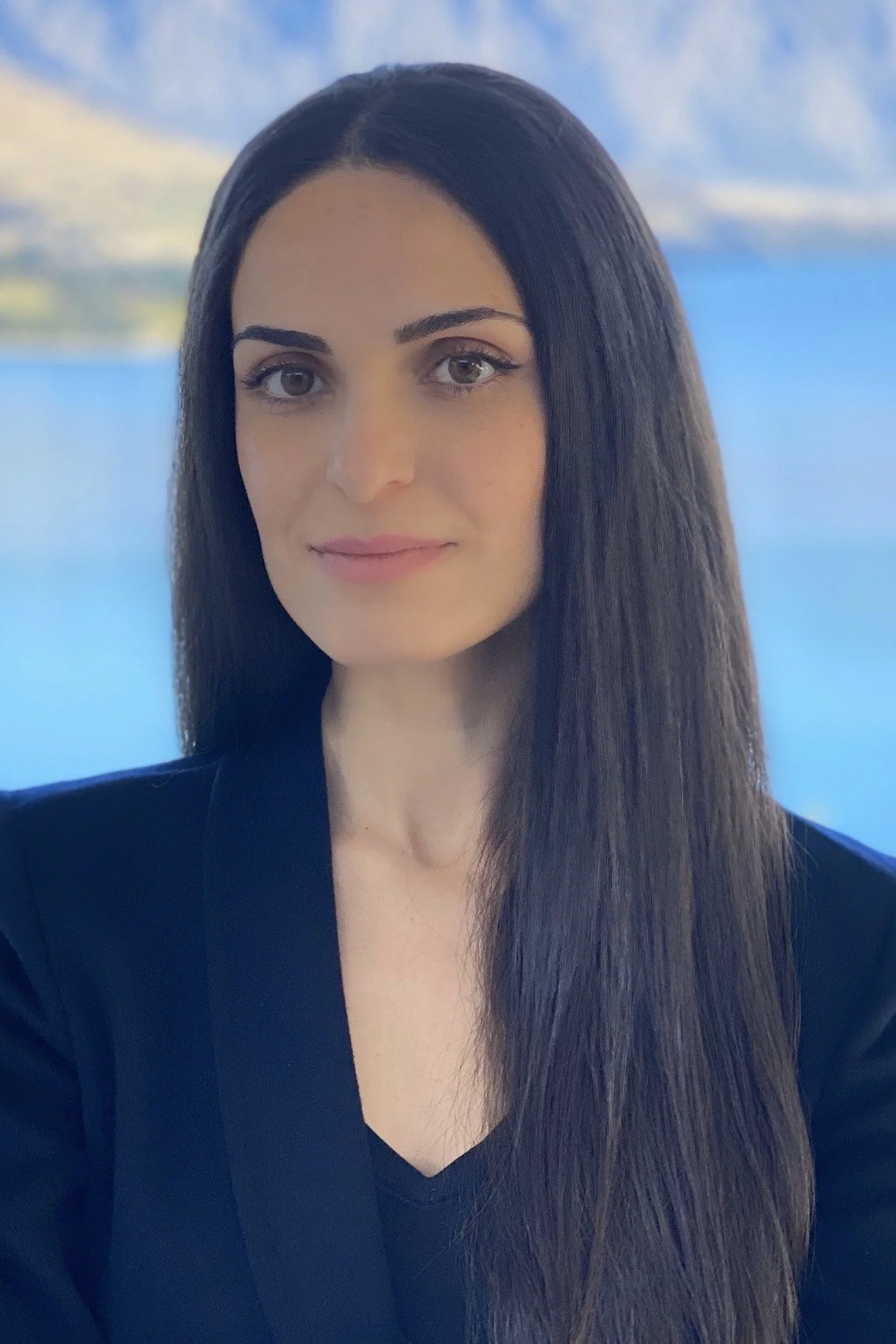 A woman with long dark hair, wearing a black top, stands in front of a blurred background of water and mountains.