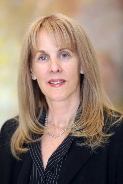 A woman with long blonde hair, wearing a black blazer and a silver necklace, looking at the camera with a neutral expression.