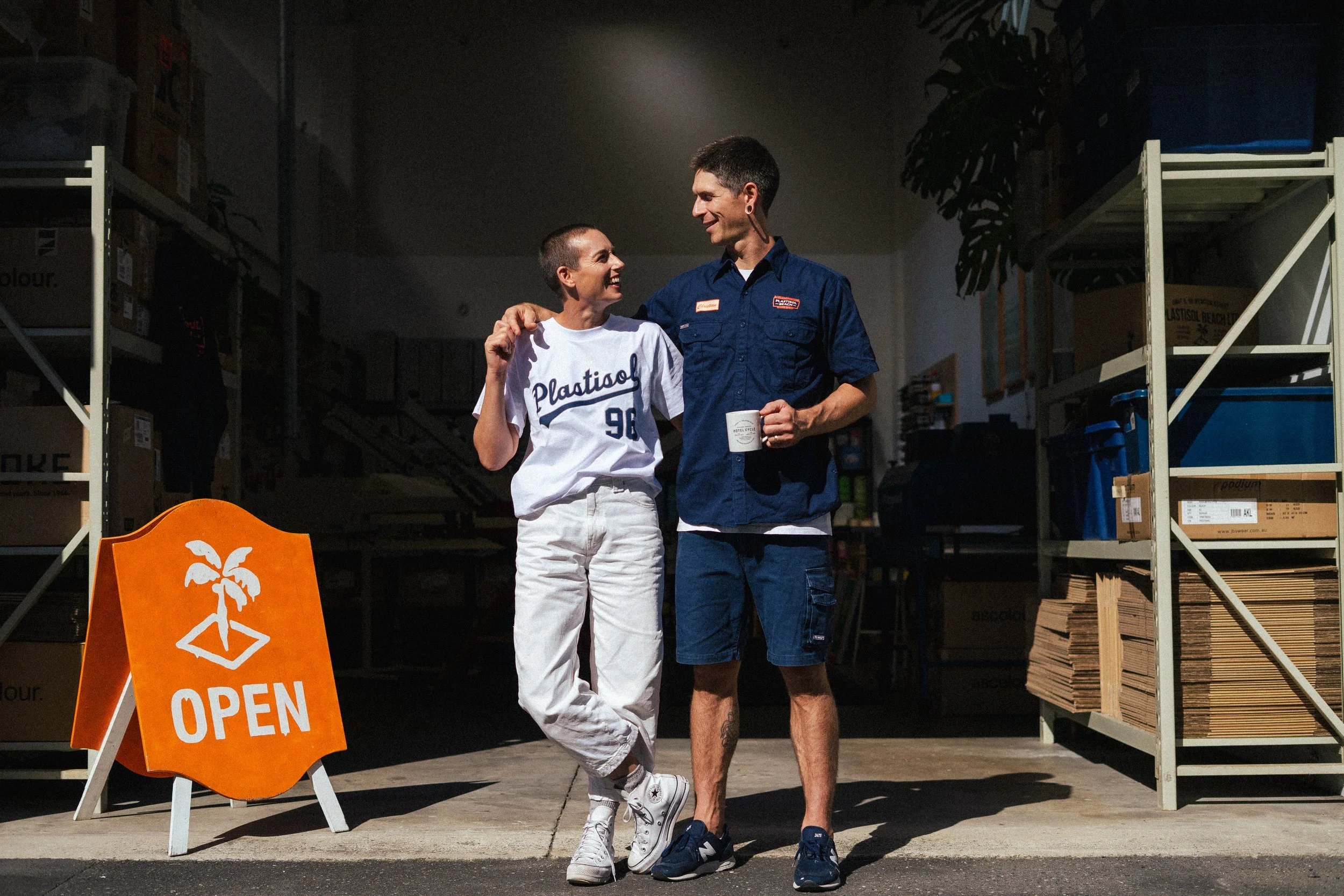 A young woman and a young man stand outside a store, smiling and looking at each other. The woman is wearing a sports jersey with the word 'Plastiso' and the number 96, white pants, and sneakers. The man is wearing a blue work shirt, shorts, and sneakers, holding a mug. An orange 'Open' sign with a palm tree logo is on the sidewalk, and store shelves with boxes are visible in the background.