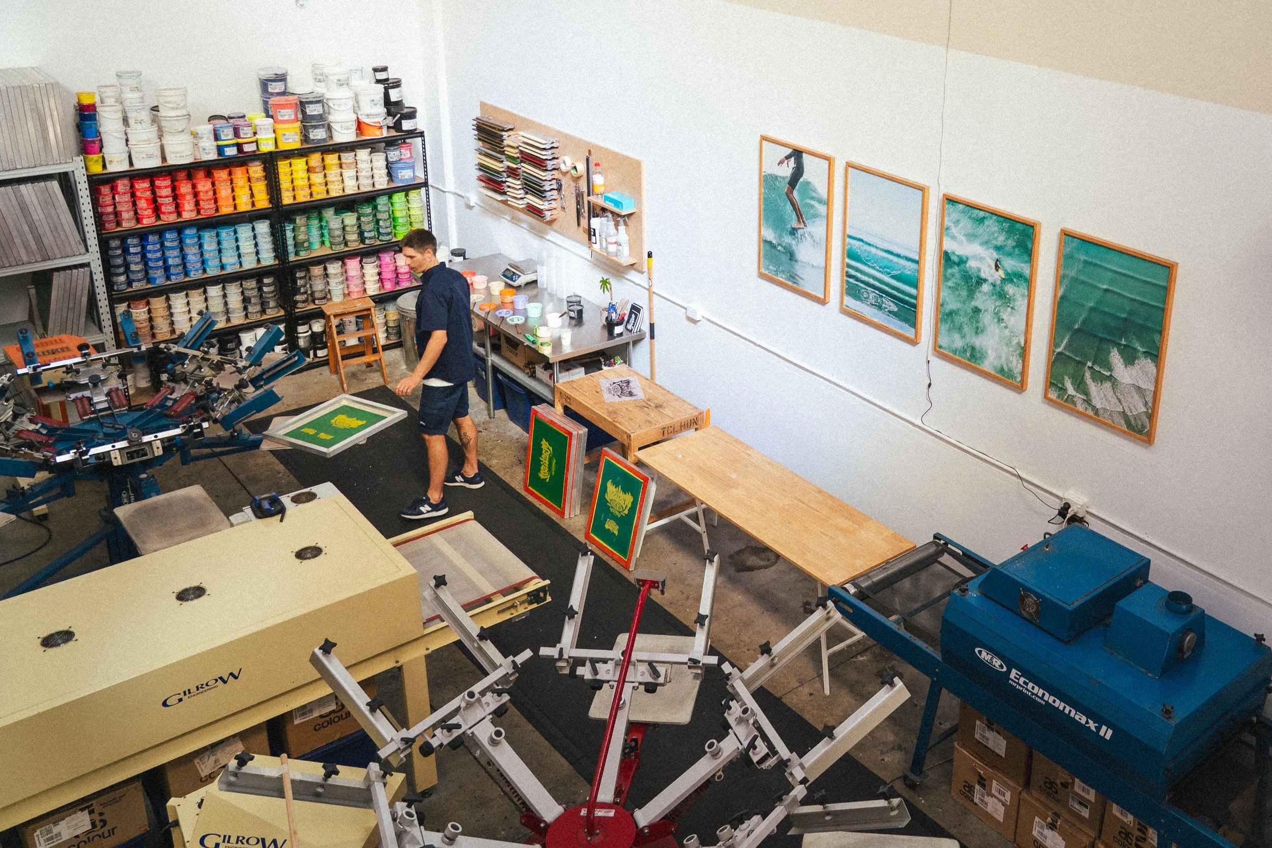 A man in a workshop surrounded by shelves of paint cans, printing presses, framed ocean surfing photographs, and various printing equipment.