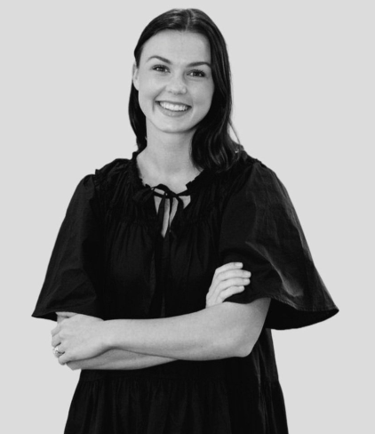 Black and white photo of a woman smiling with her arms crossed, wearing a dark dress with puffed sleeves and a tie at the neckline.