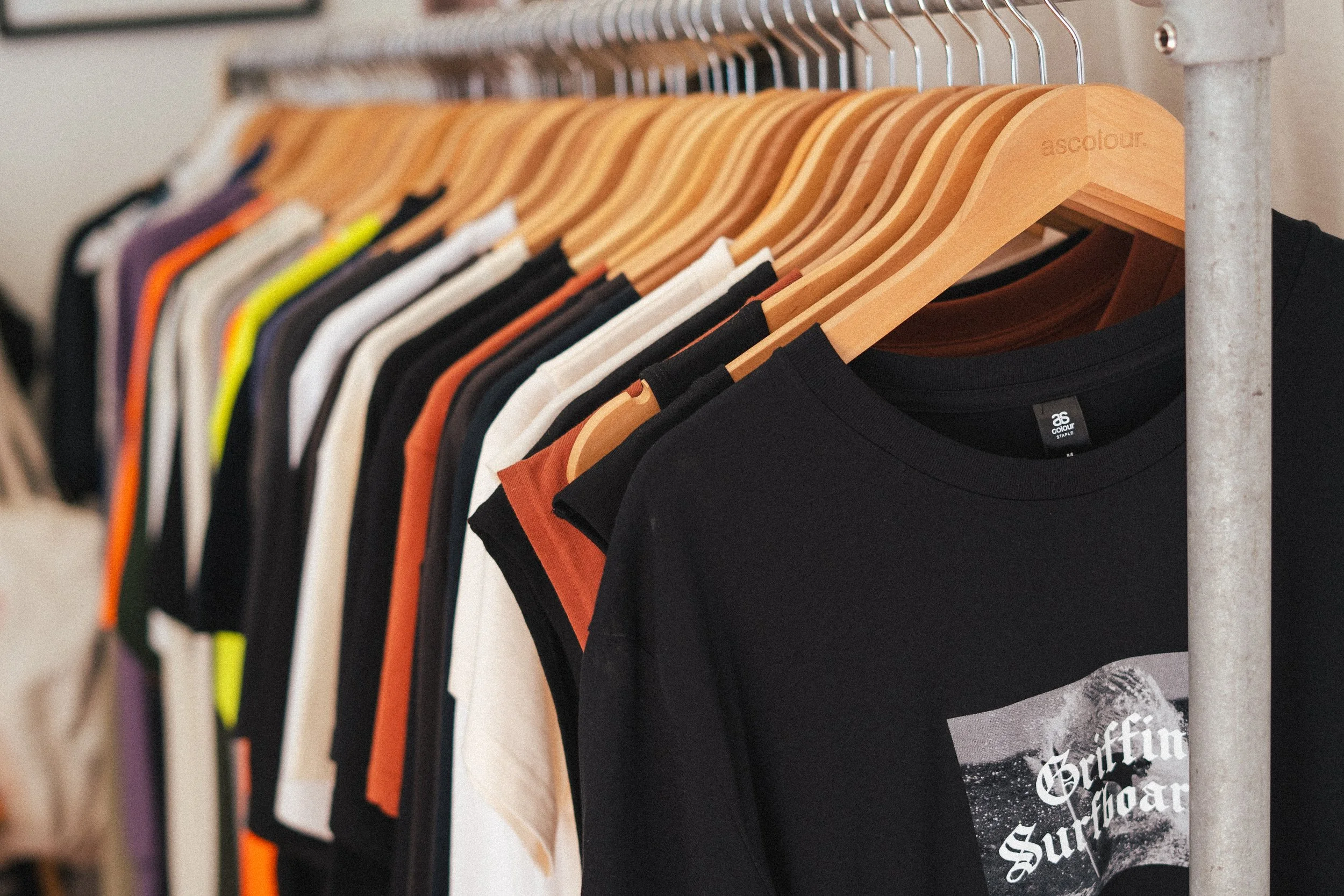 A clothing rack with various T-shirts on wooden hangers, including a prominently visible black T-shirt with white text and graphic.
