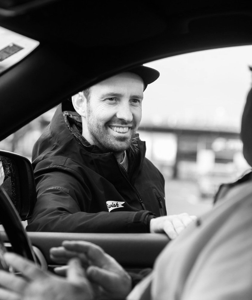 A smiling man wearing a jacket and cap, standing outside a car, interacting with someone inside the vehicle.