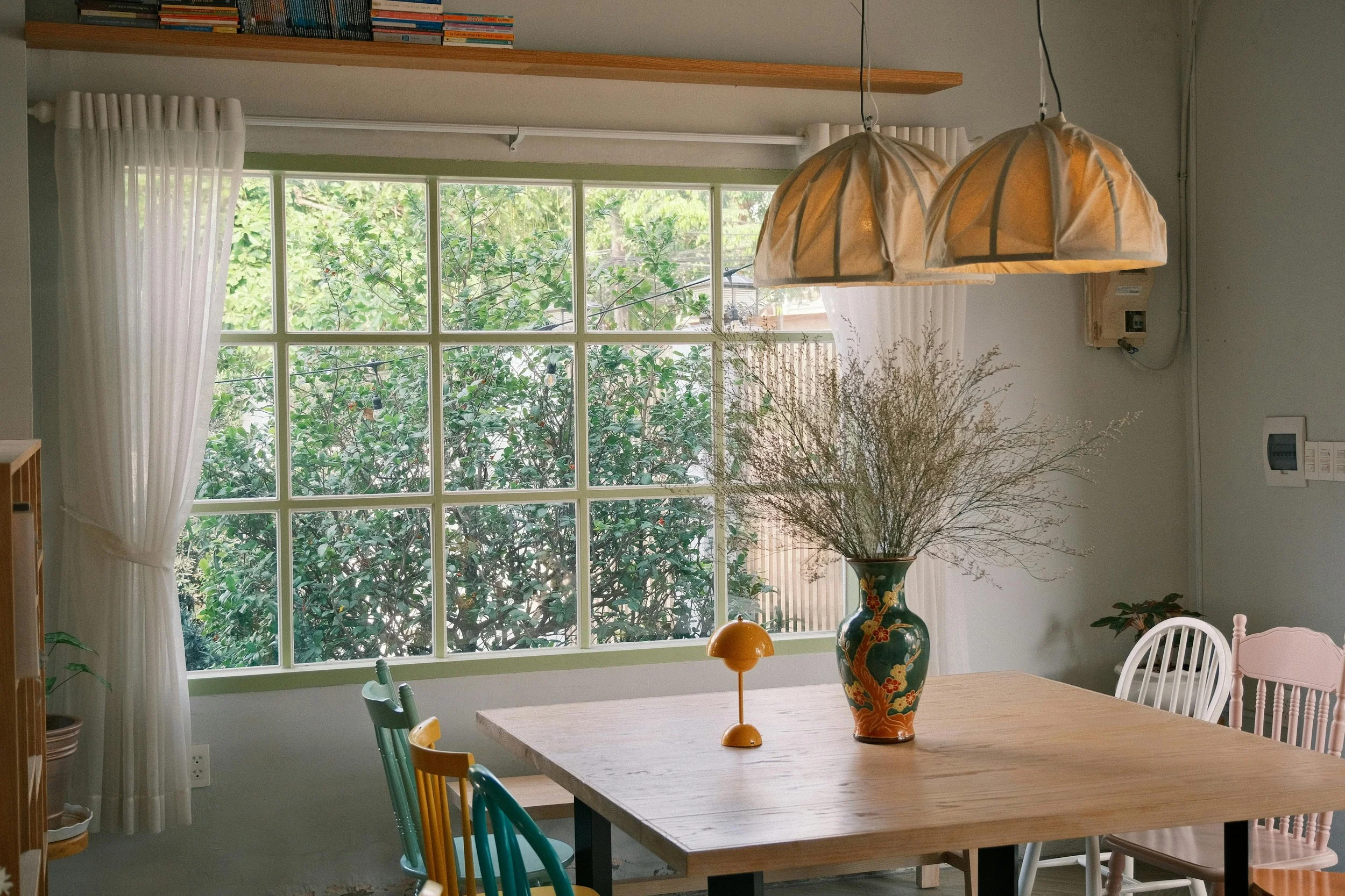 A dining room with a wooden table, a large window showing greenery outside, and a large vase with branches on the table. Two pendant lights hang above the table, and there are colorful chairs around it.