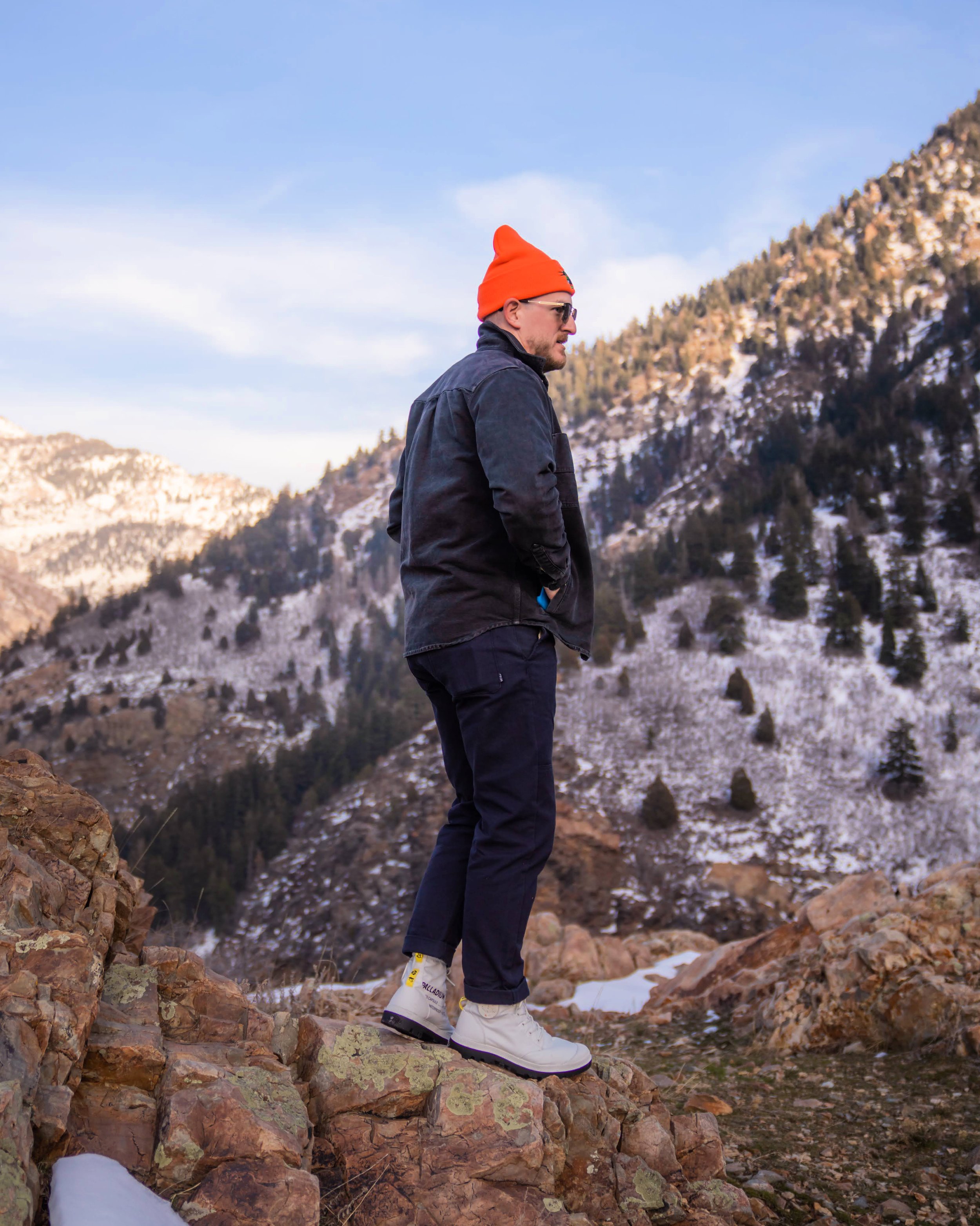 Man standing on rocky terrain in a mountainous area during winter, wearing a black jacket, navy pants, white boots, an orange beanie, and sunglasses.