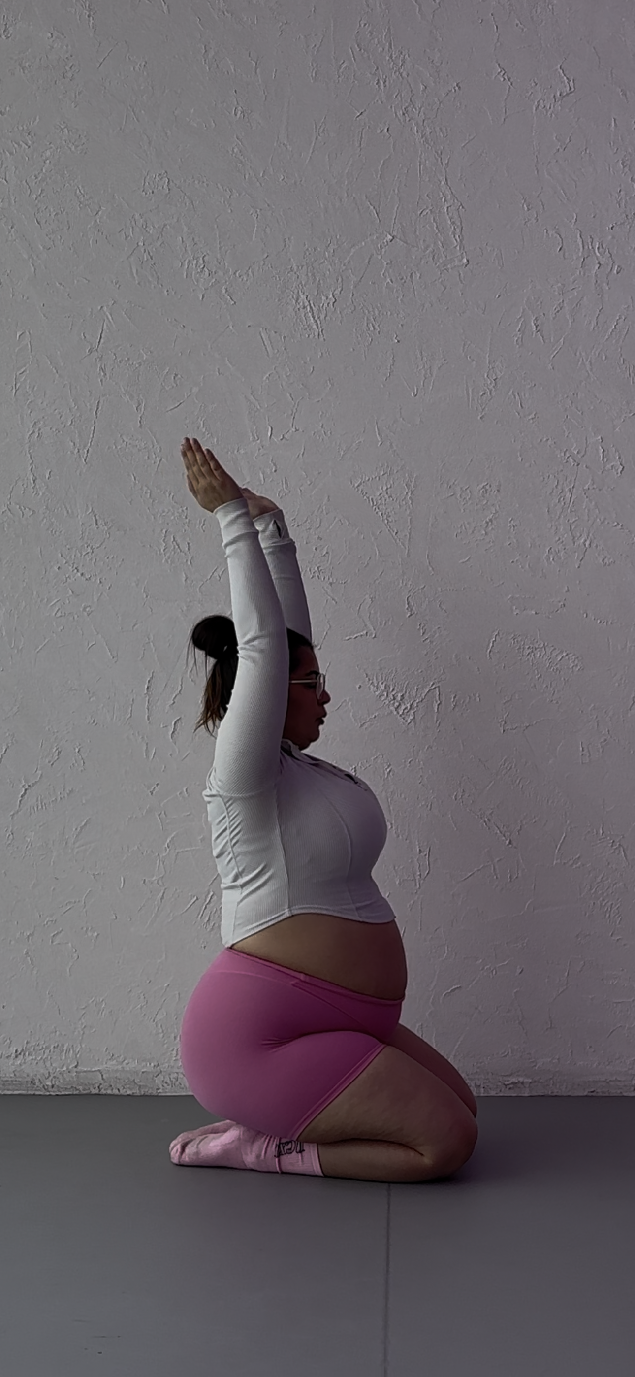 A woman with glasses and a ponytail is doing a yoga pose on a gray floor against a textured white wall. She is wearing a white zip-up top, pink shorts, and pink socks, with one arm raised upward and the other resting on her knee.