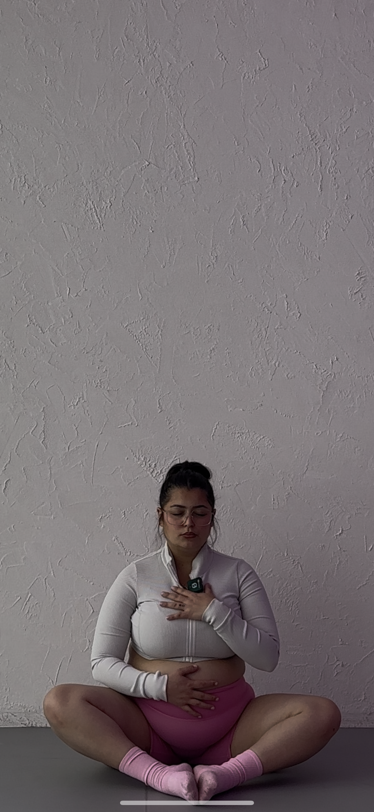 A woman sitting cross-legged on the floor meditating in front of a textured white wall, wearing a white zip-up jacket, pink shorts, pink socks, and glasses.