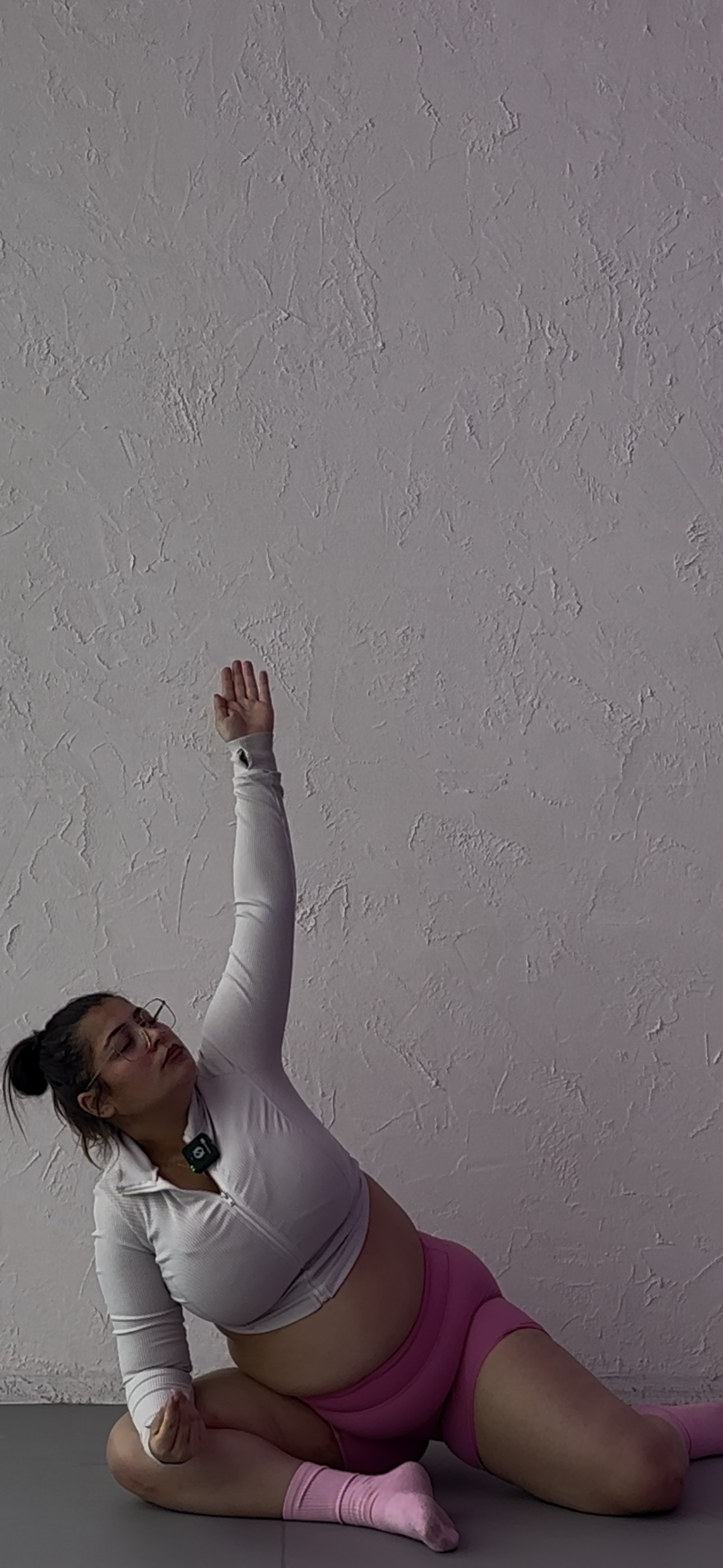 Woman sitting cross-legged on the floor in front of a textured white wall, wearing glasses, a half-zip white top, pink shorts, and pink socks, with her right hand on her chest and left hand on her stomach, eyes closed.