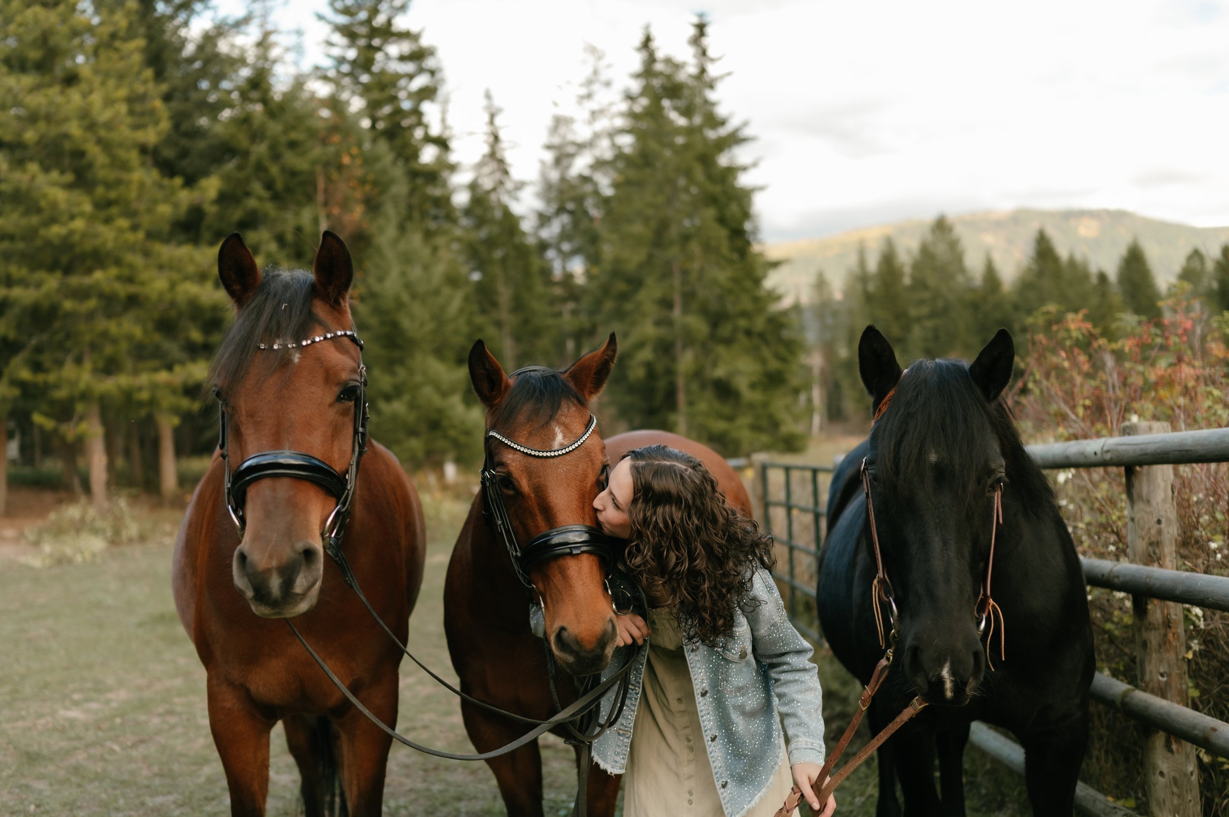 A person with curly hair in a denim jacket is kissing one of three horses in a woodland area. The horses, two brown and one black, are wearing bridles and standing beside a wooden fence surrounded by trees.