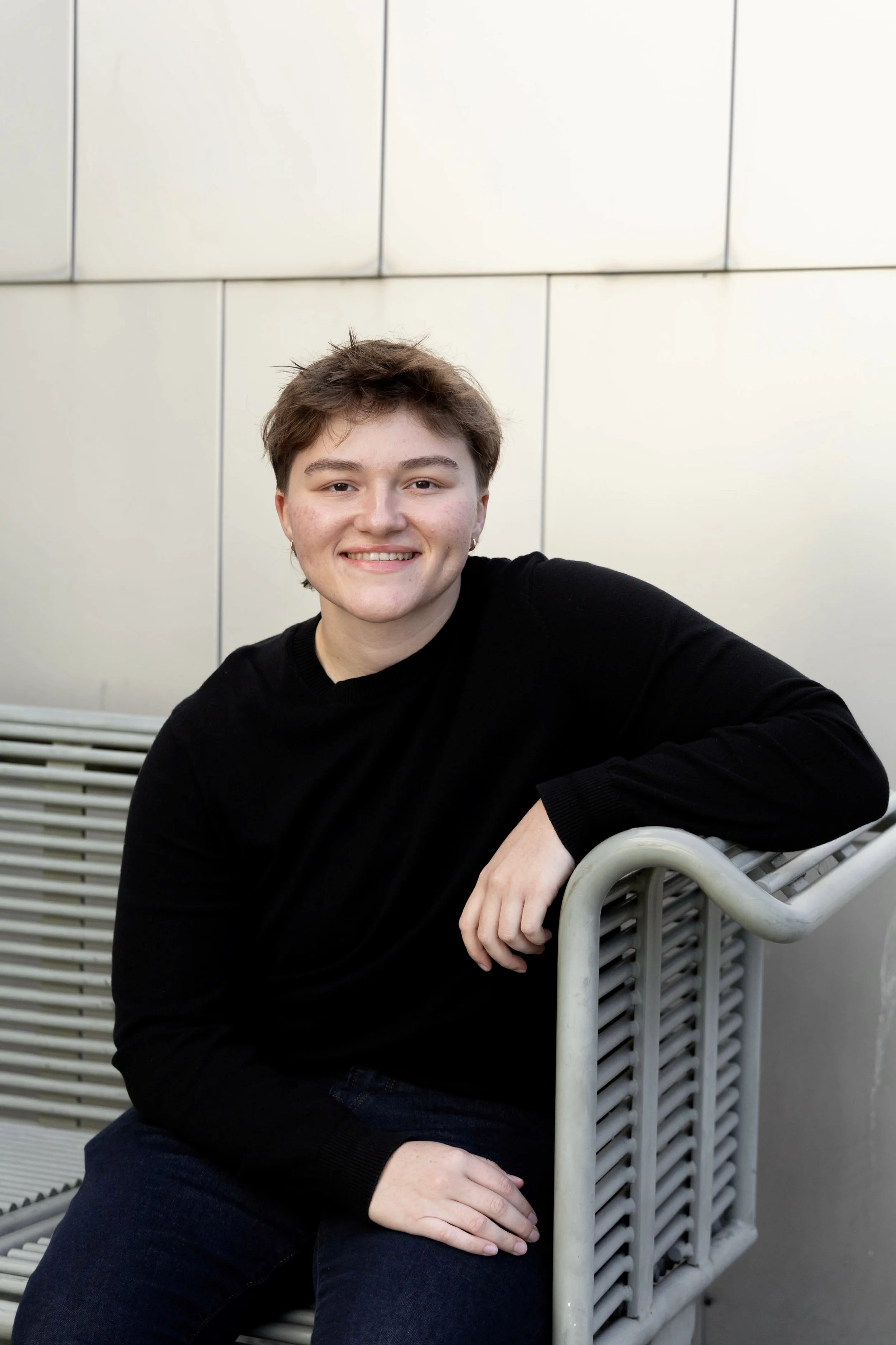 A smiling person with short brown hair sitting on a metal bench outdoors against a beige wall.