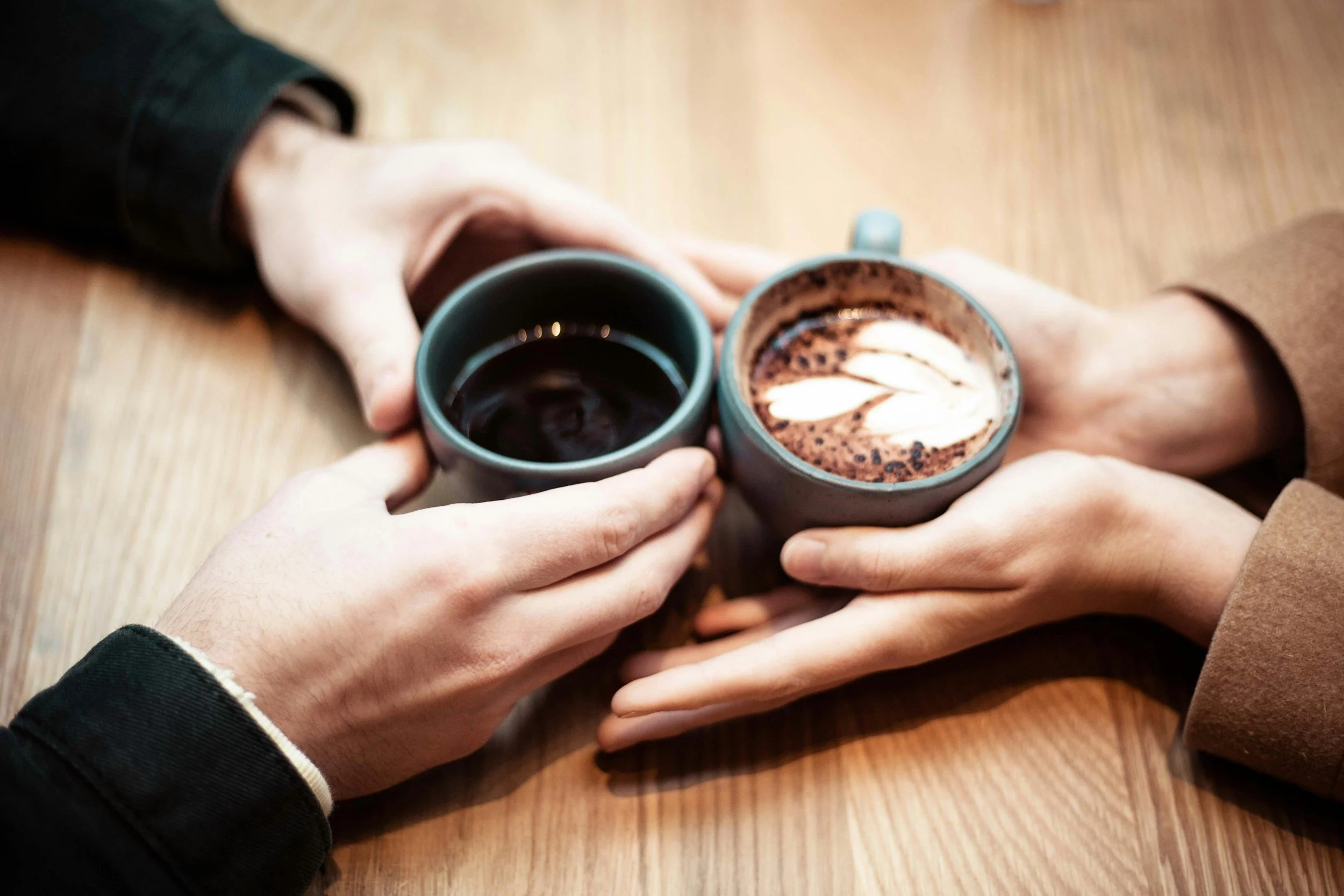 Two people holding ceramic cups of coffee at a wooden table, one with black coffee and the other with a latte topped with latte art.