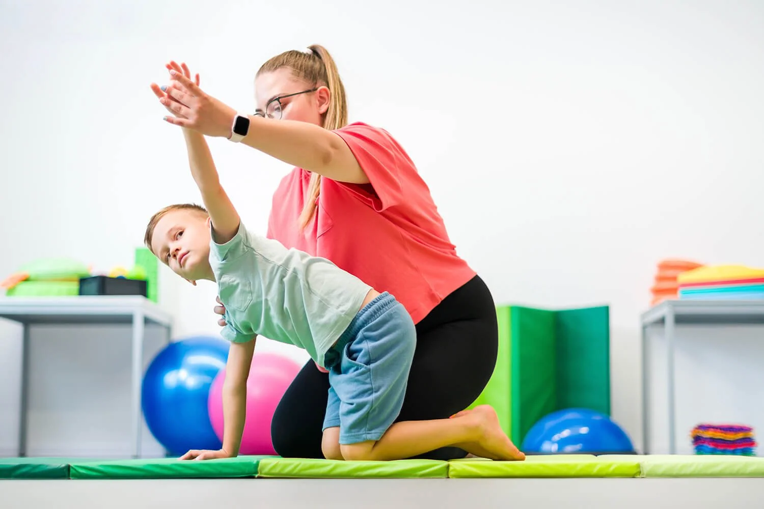 A physical therapist assisting a young boy with a yoga exercise on a padded mat in a therapy or gym setting, with exercise balls and colourful equipment in the background.