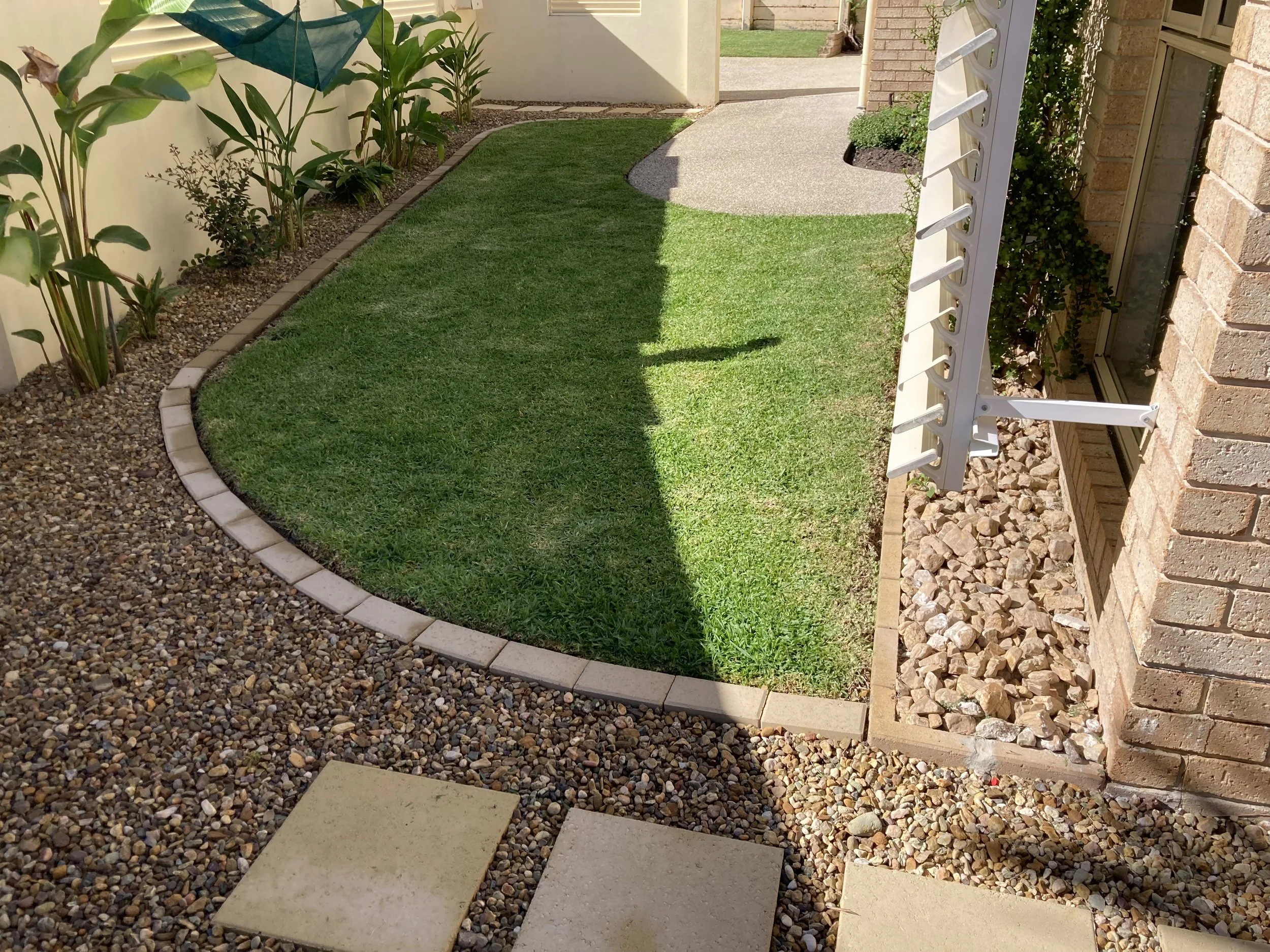 Backyard garden with a curved grass lawn bordered by small beige bricks, surrounded by gravel and stone walkway, with plants along the fence, and a white outdoor drying rack extending from the brick wall of a house.