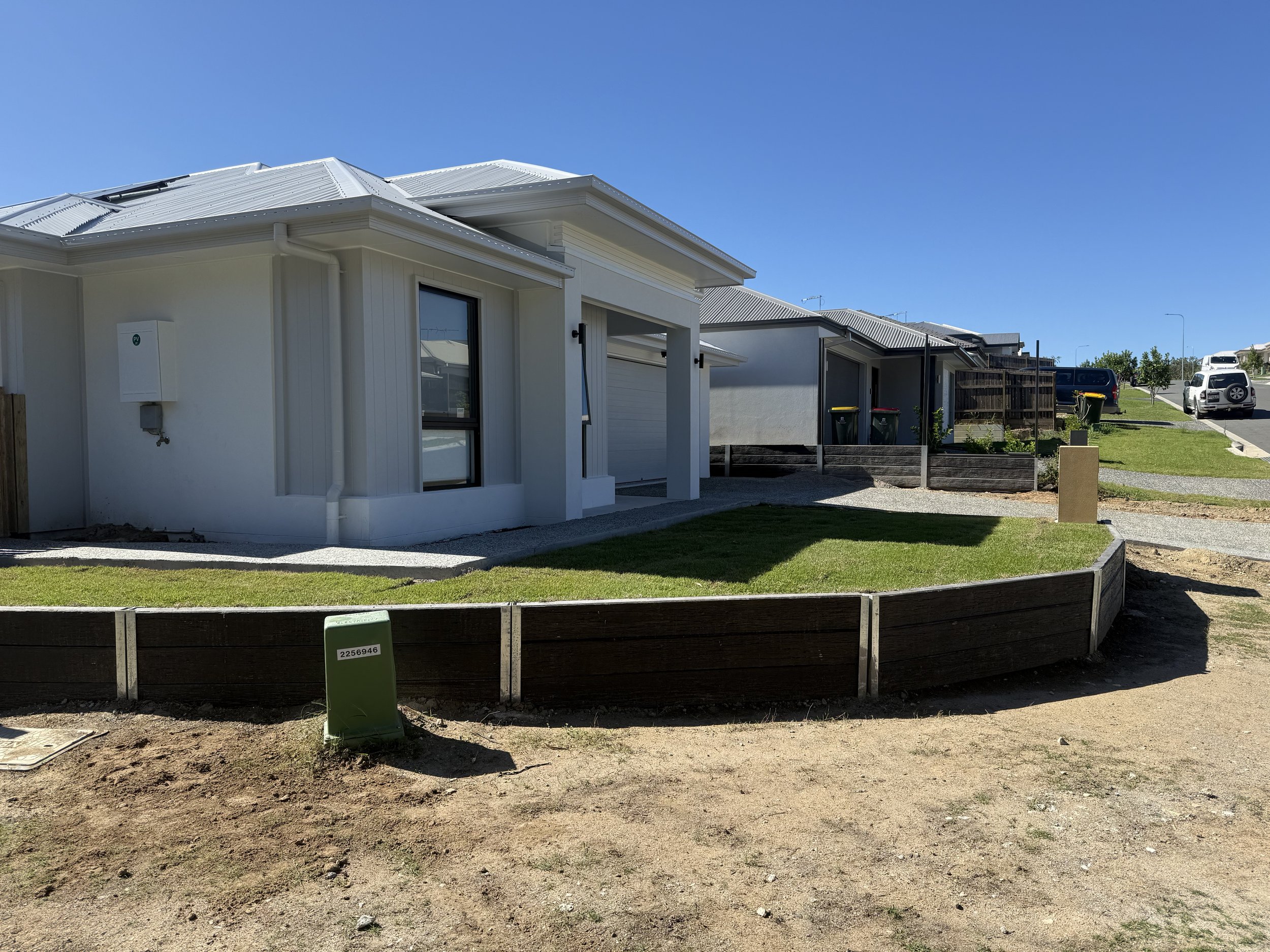 Modern white house with a metal roof, new landscaping with a small lawn and wooden border, and several parked cars in the background.