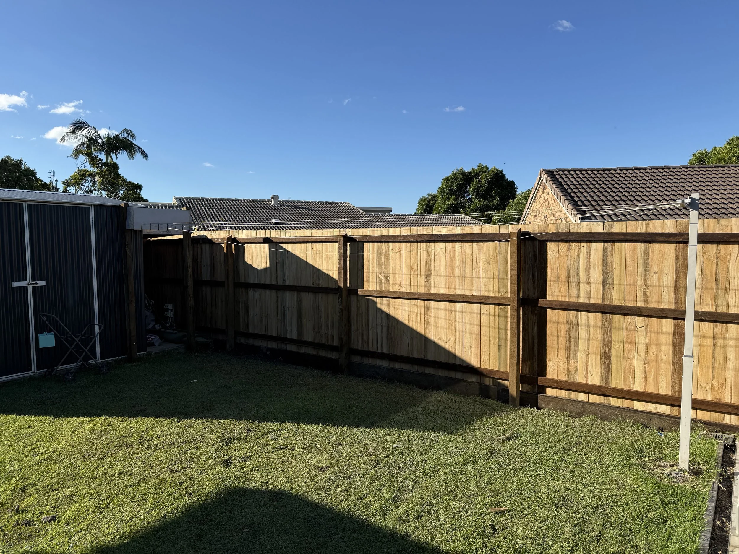 Backyard with green grass, wooden privacy fence, metal shed, laundry pole, and neighboring rooftops with cloudy blue sky.