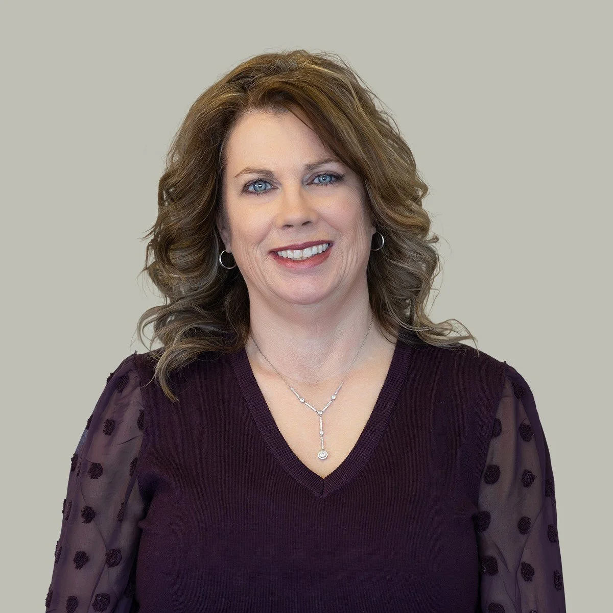 A woman with shoulder-length wavy brown hair and blue eyes, smiling, wearing a dark purple sweater with sheer polka-dot sleeves, silver hoop earrings, and layered silver necklaces against a plain light gray background.