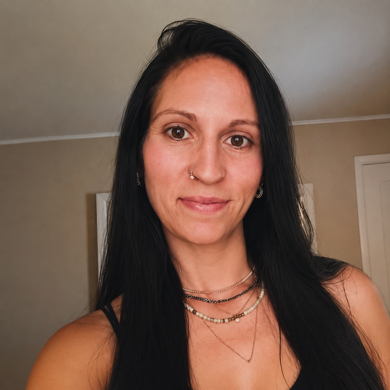 A woman with long black hair, brown eyes, and light skin, wearing multiple layered necklaces and earrings, smiling softly indoors against a beige wall with white door frames in the background.