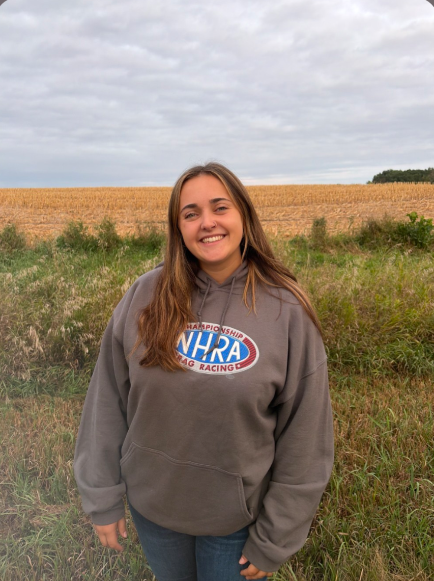 Young woman with long brown hair smiling outdoors in a field with green grass and a harvested crop in the background under a cloudy sky, wearing a gray hoodie with the NHRA logo.