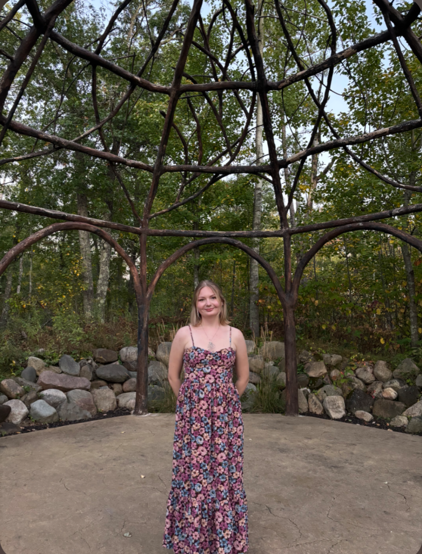 Young woman in a floral dress standing outdoors on a cracked concrete surface, with a decorative wooden arch structure and trees with green and yellow leaves in the background.