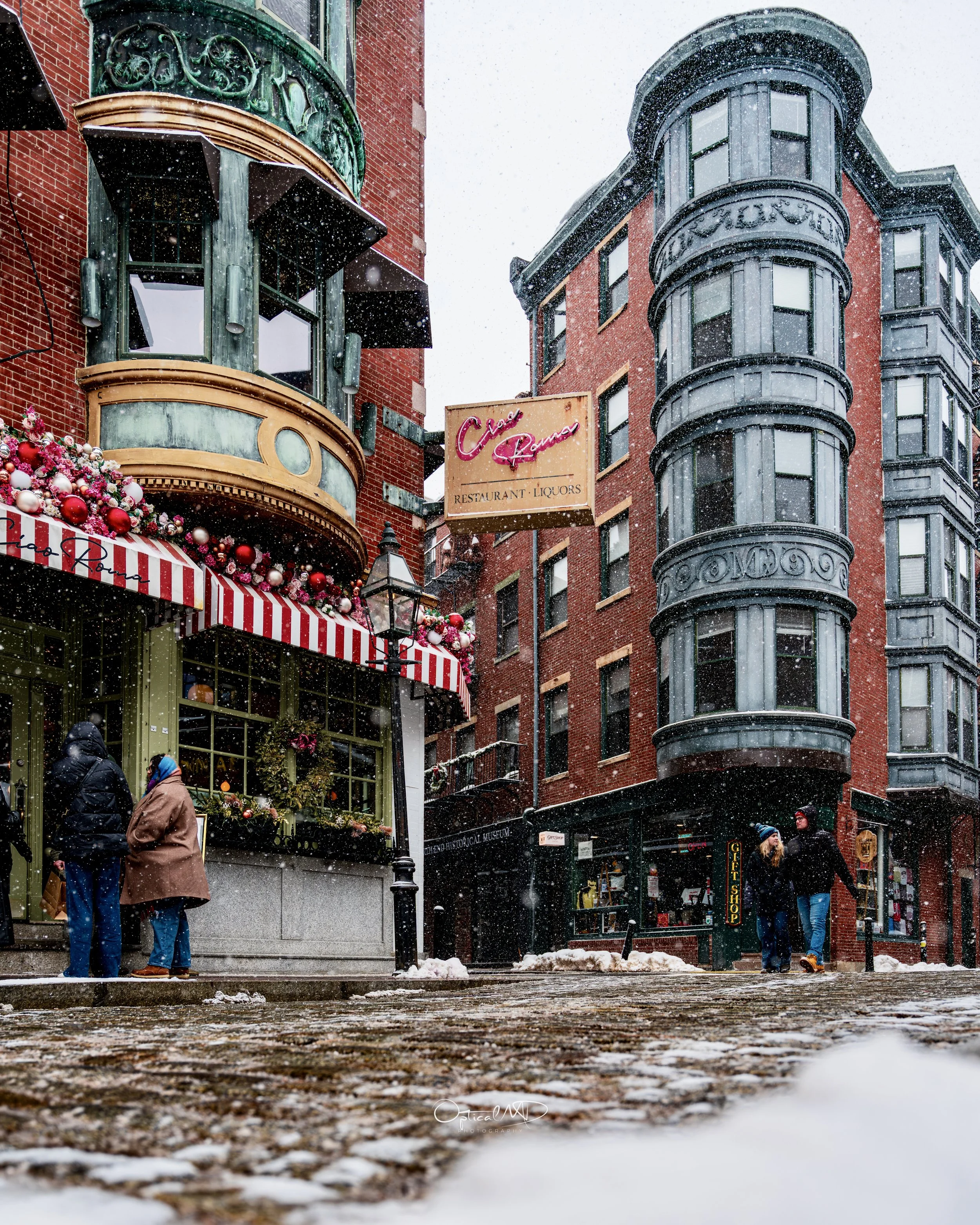 Snow falling on a street with people walking, red brick buildings, a restaurant with holiday decorations, and a vintage-style lamppost.