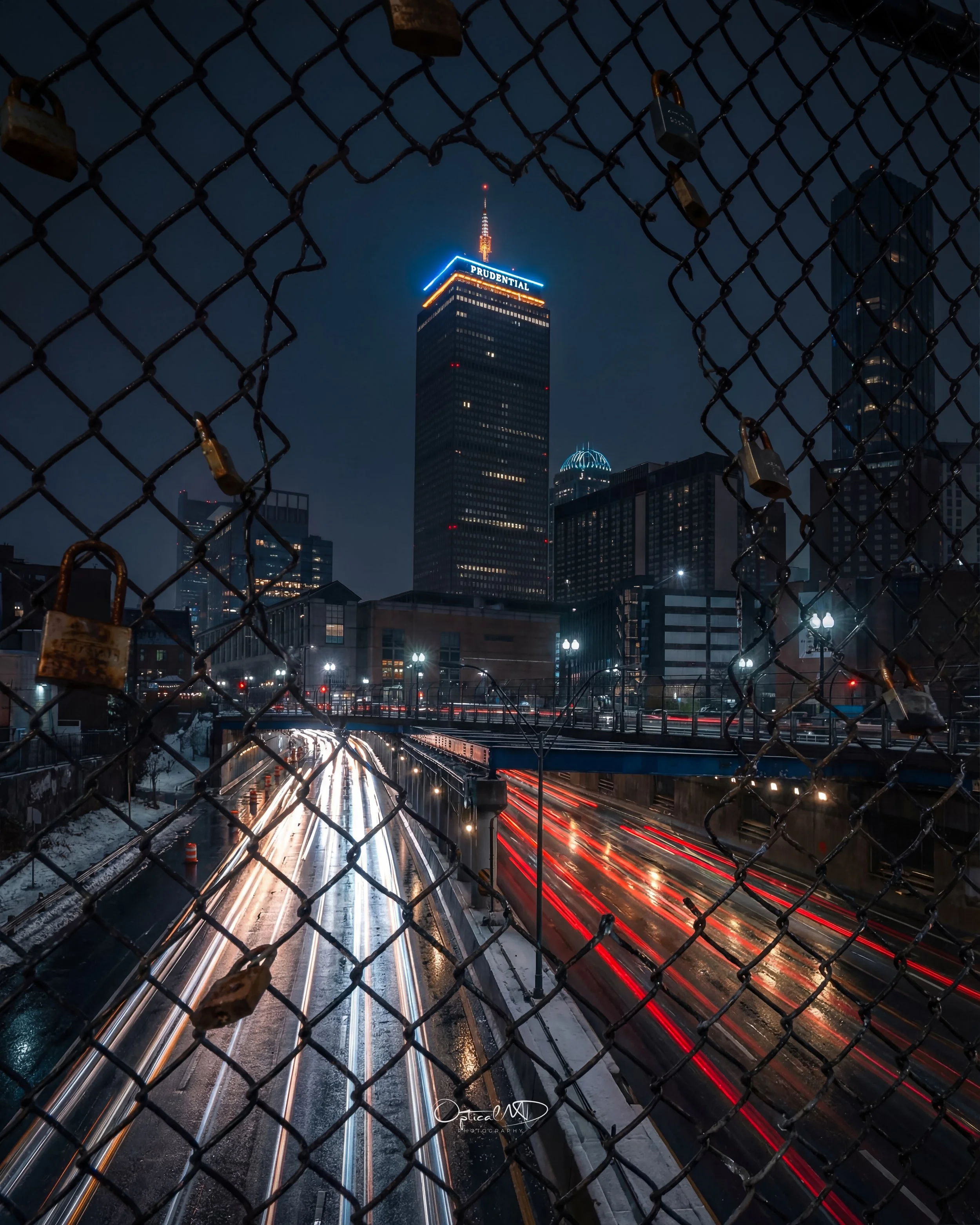 Night view of a city skyline with the Prudential Tower, seen through a chain-link fence with padlocks, and streaks of moving car lights on a wet highway.