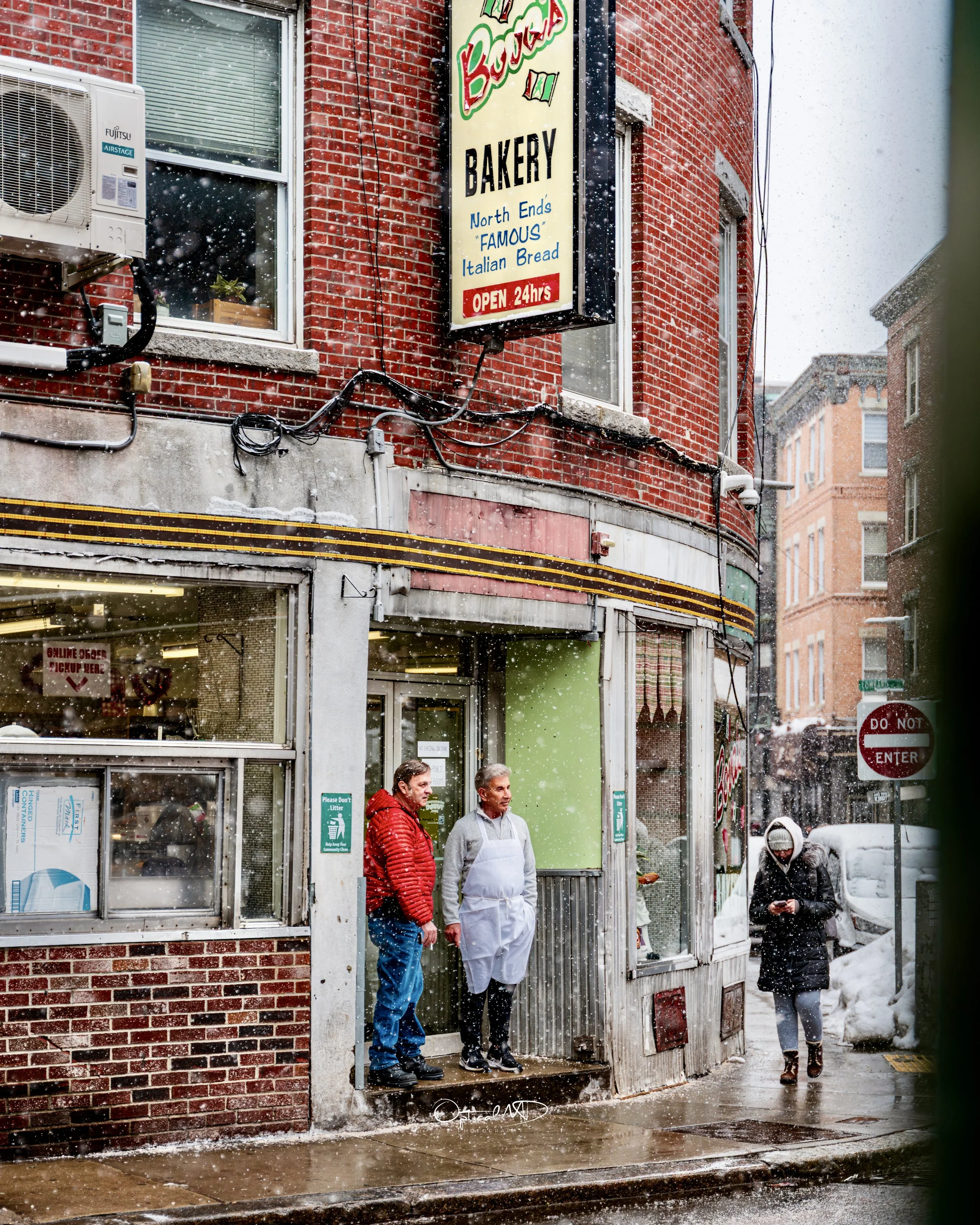 People standing outside a bakery on a snowy day. Two men are talking near the entrance, one of them wearing a white apron. A woman wearing a mask and coat walks by on the sidewalk.