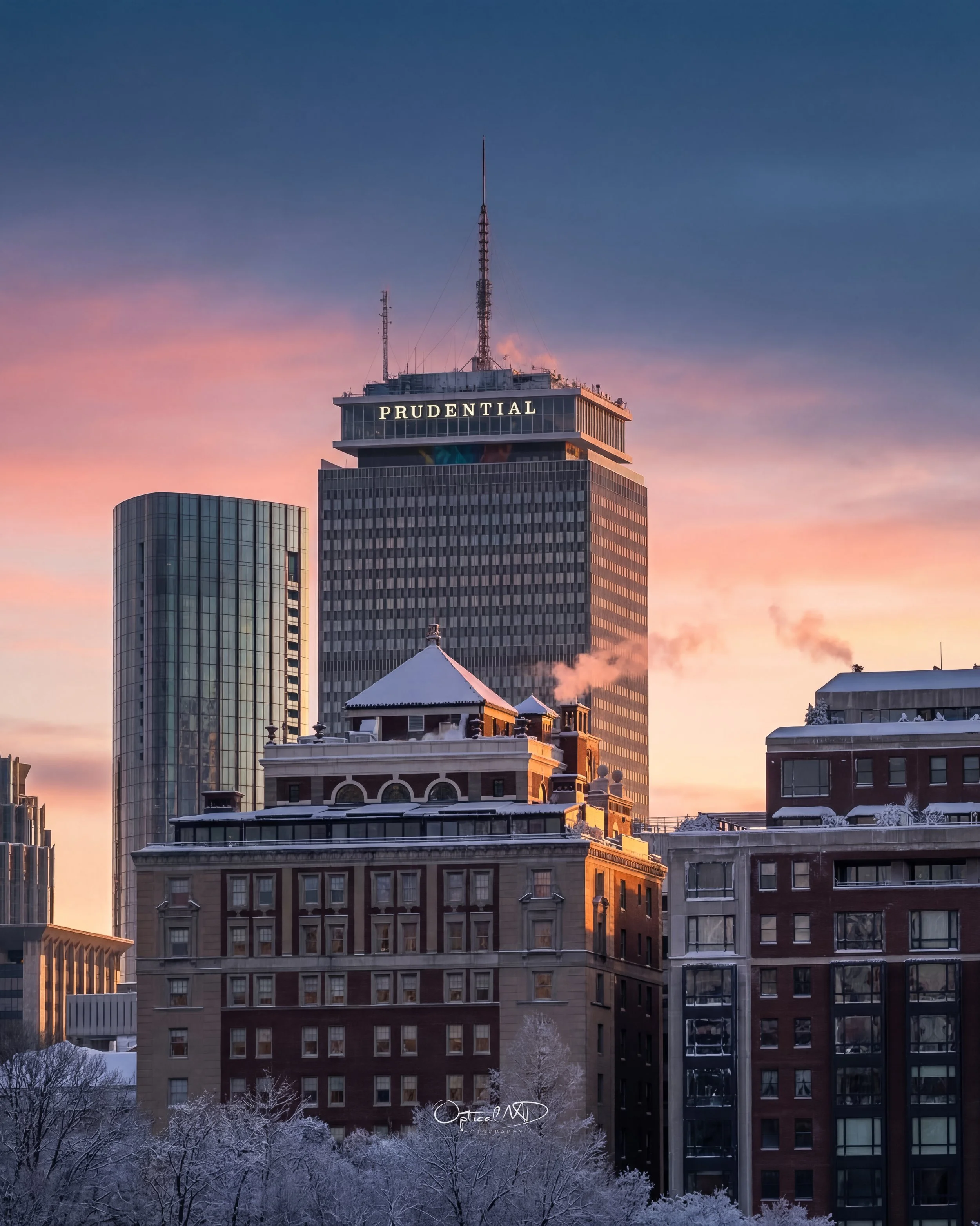City skyscrapers at sunset with snow-covered trees in foreground, including Prudential Tower with illuminated sign.