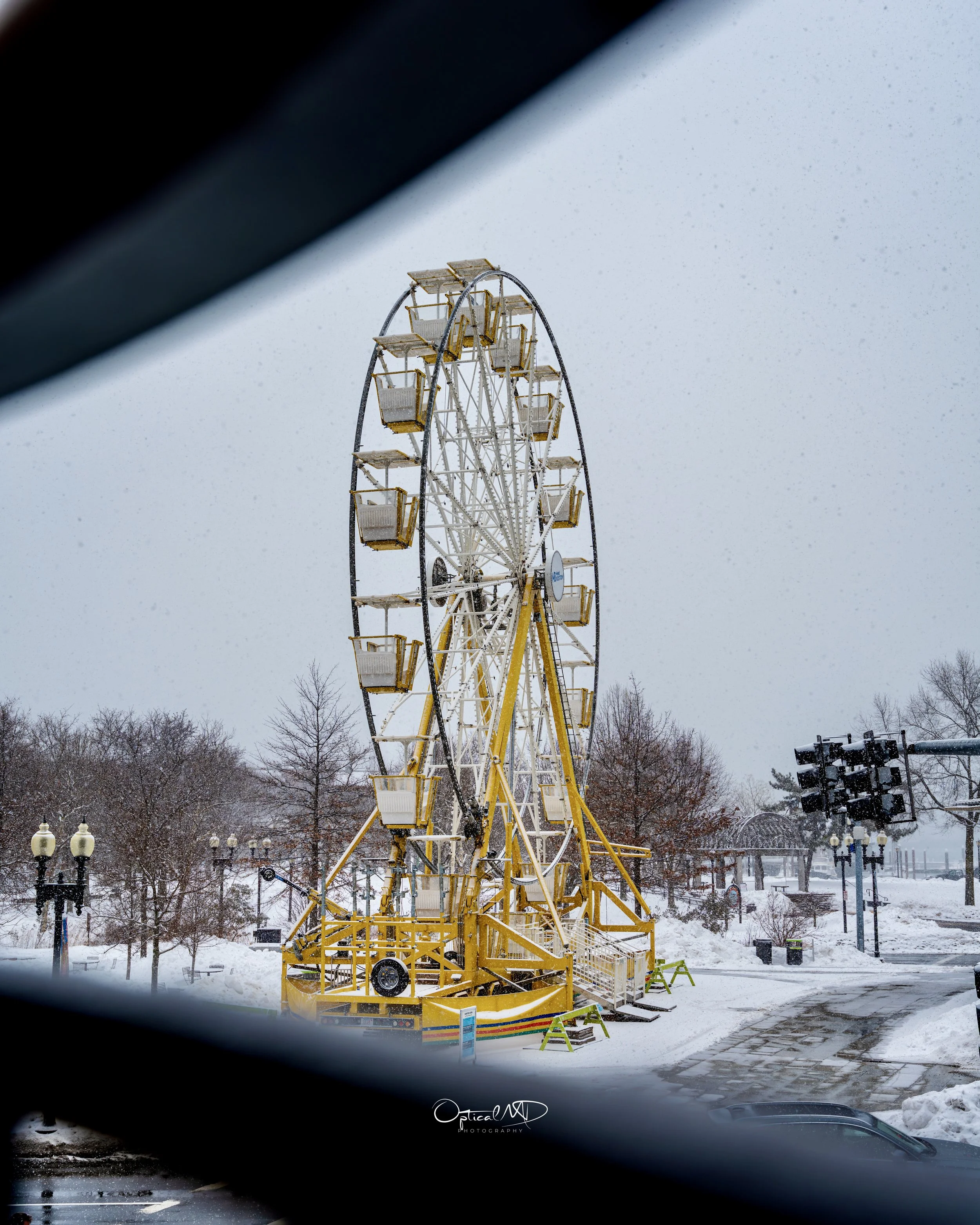 Ferris wheel in a snow-covered park viewed through a car window.