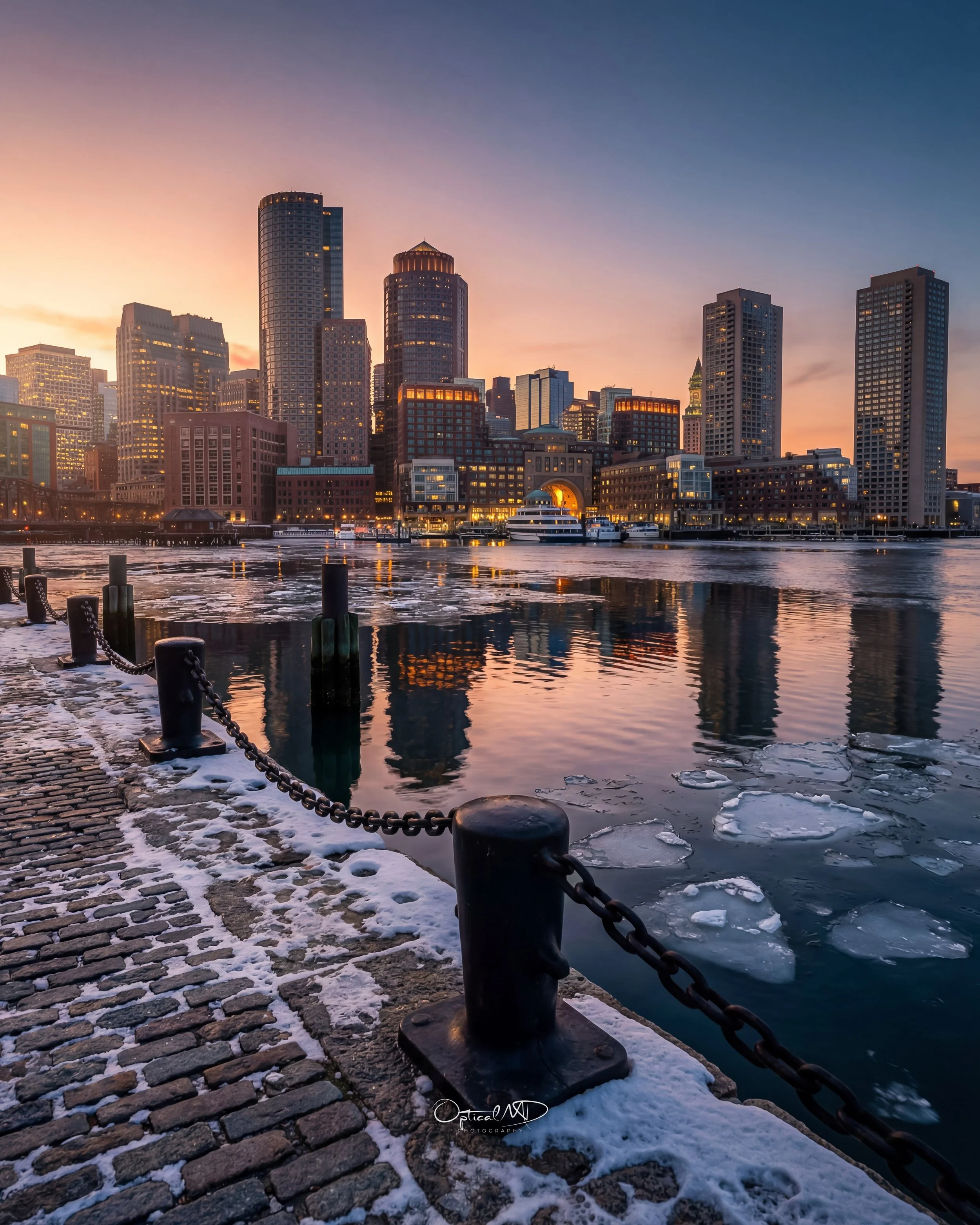 City skyline with tall buildings at sunset, reflecting on the water with ice and snow on the dock and chain barrier in the foreground.