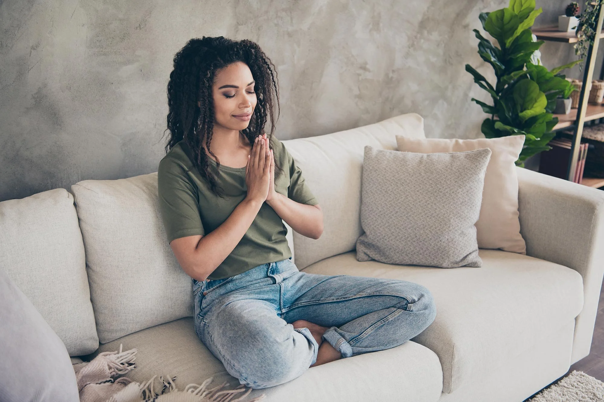 A woman sitting cross-legged on a beige sofa with her eyes closed, hands pressed together in a prayer position, practicing meditation or mindfulness in a cozy living room.