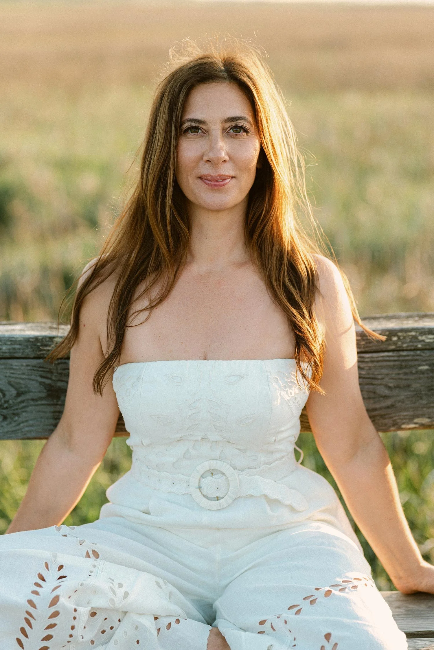 A woman with long brown hair wearing a white strapless dress sitting on a wooden bench outdoors during golden hour.