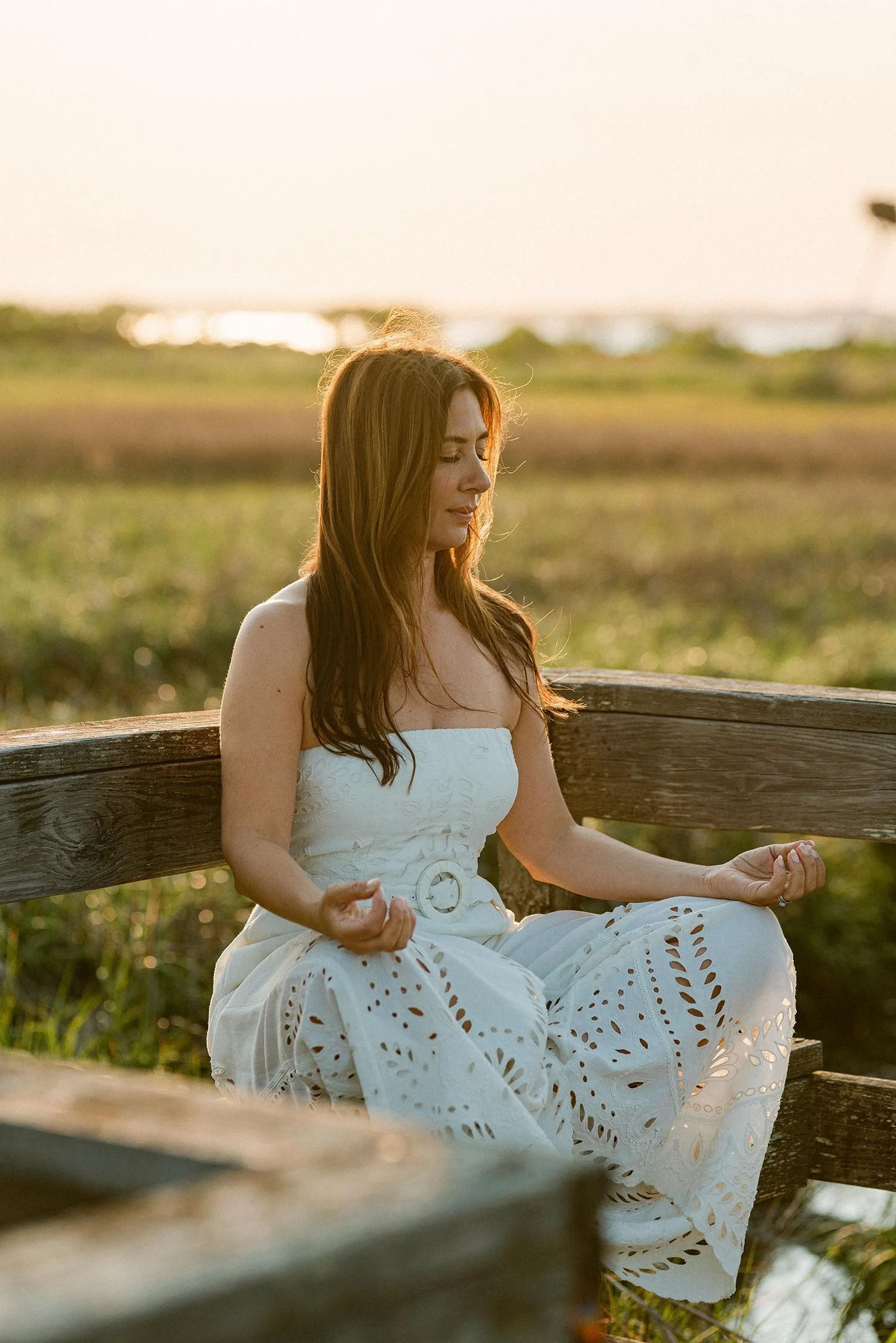A woman with long, red hair dressed in a white, strapless dress sitting cross-legged on a wooden bench outdoors at sunset, practicing meditation with her eyes closed.