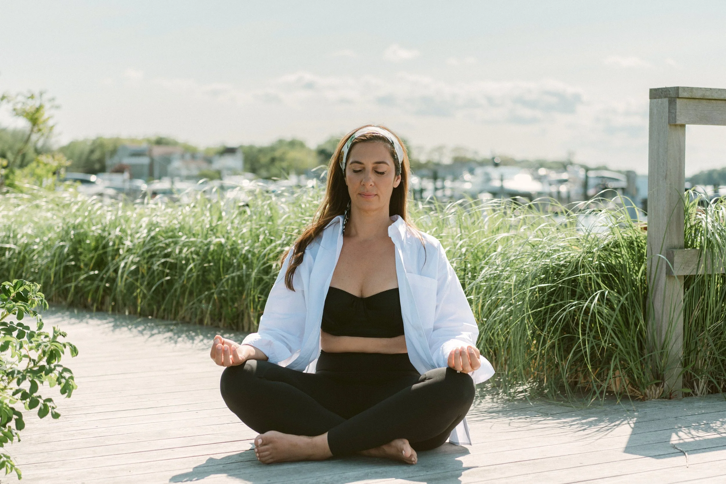 Woman practicing meditation outdoors sitting cross-legged on a wooden pathway with her hands on her knees, eyes closed, surrounded by green plants, with a cloudy sky in the background.
