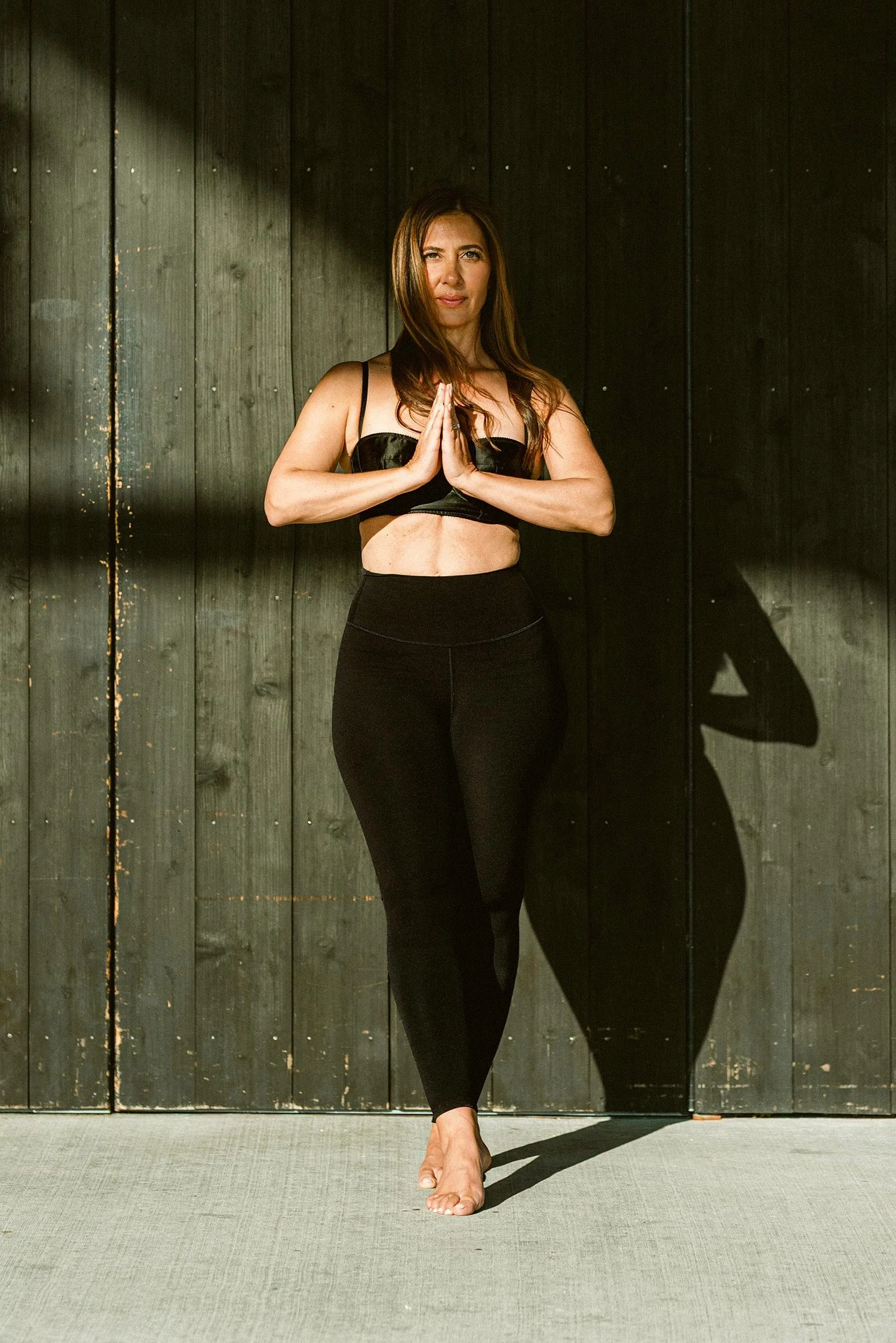 A woman practicing yoga in a yoga studio, standing on one foot with her hands pressed together in front of her chest in a prayer position, wearing black athletic wear, with sunlight and shadows on a black wood-paneled wall.