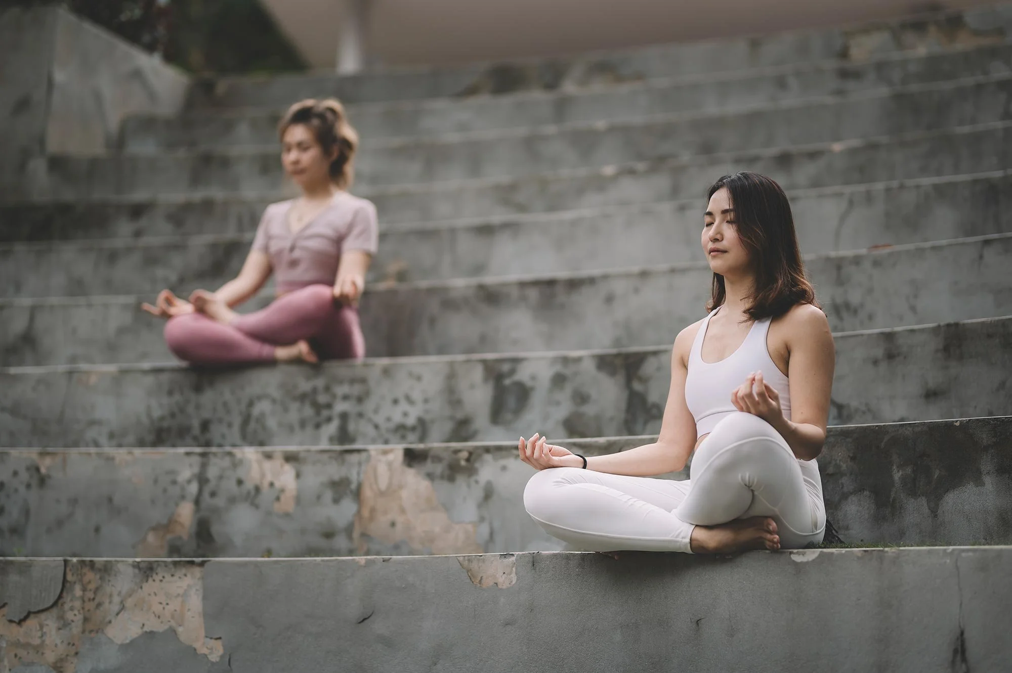 Two women practicing meditation on concrete stairs, sitting cross-legged with eyes closed, one in a white tank top and the other in a pink outfit.
