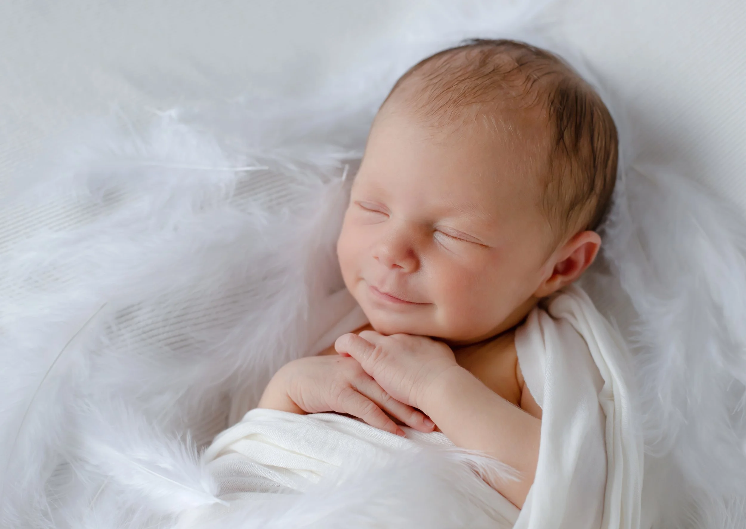 Close-up photo of a sleeping newborn baby with closed eyes, lying on a white furry blanket, wearing a white outfit.