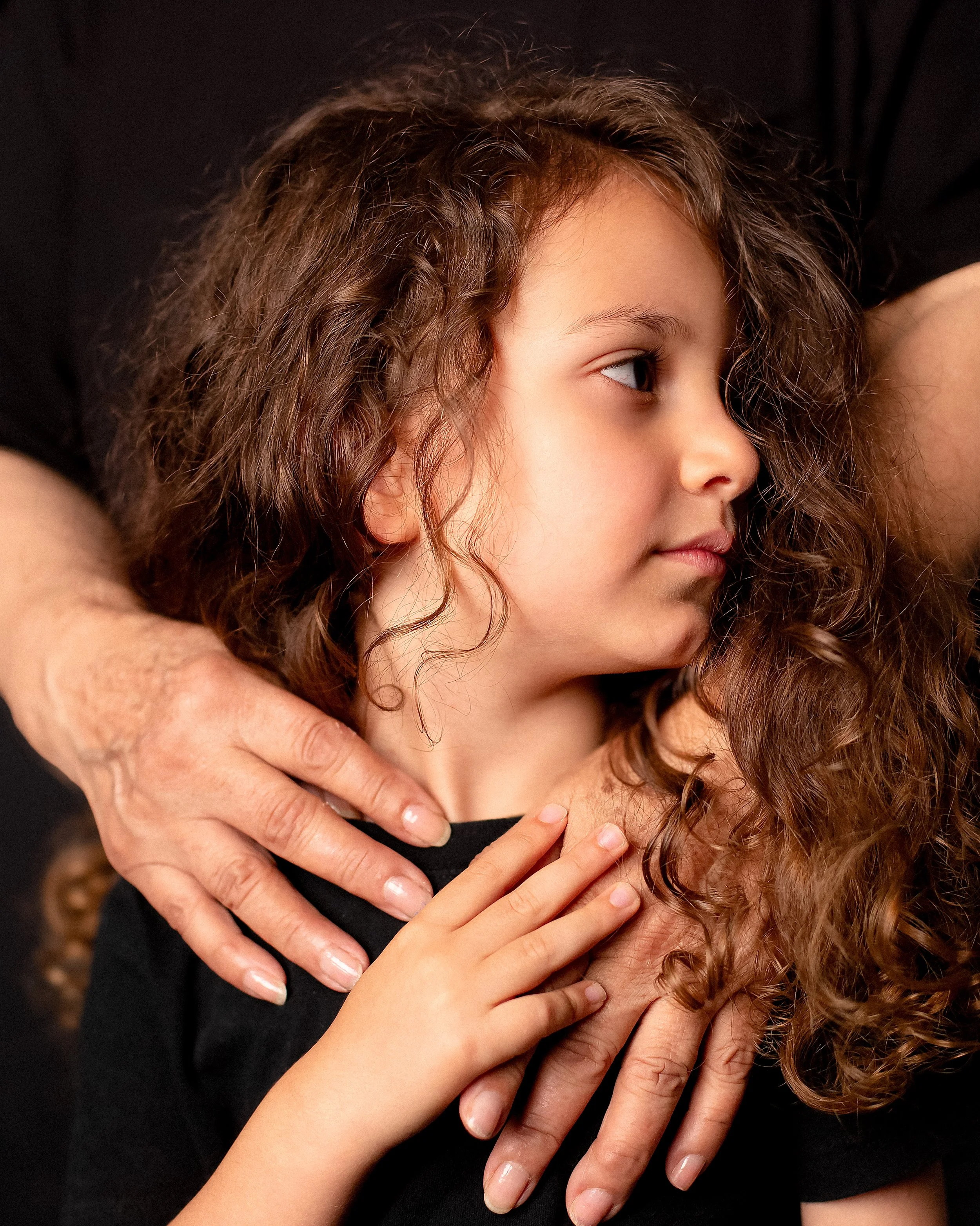 Close-up of a young girl with curly hair being hugged by an adult with multiple hands gently holding her around the shoulders and chest against a black background.